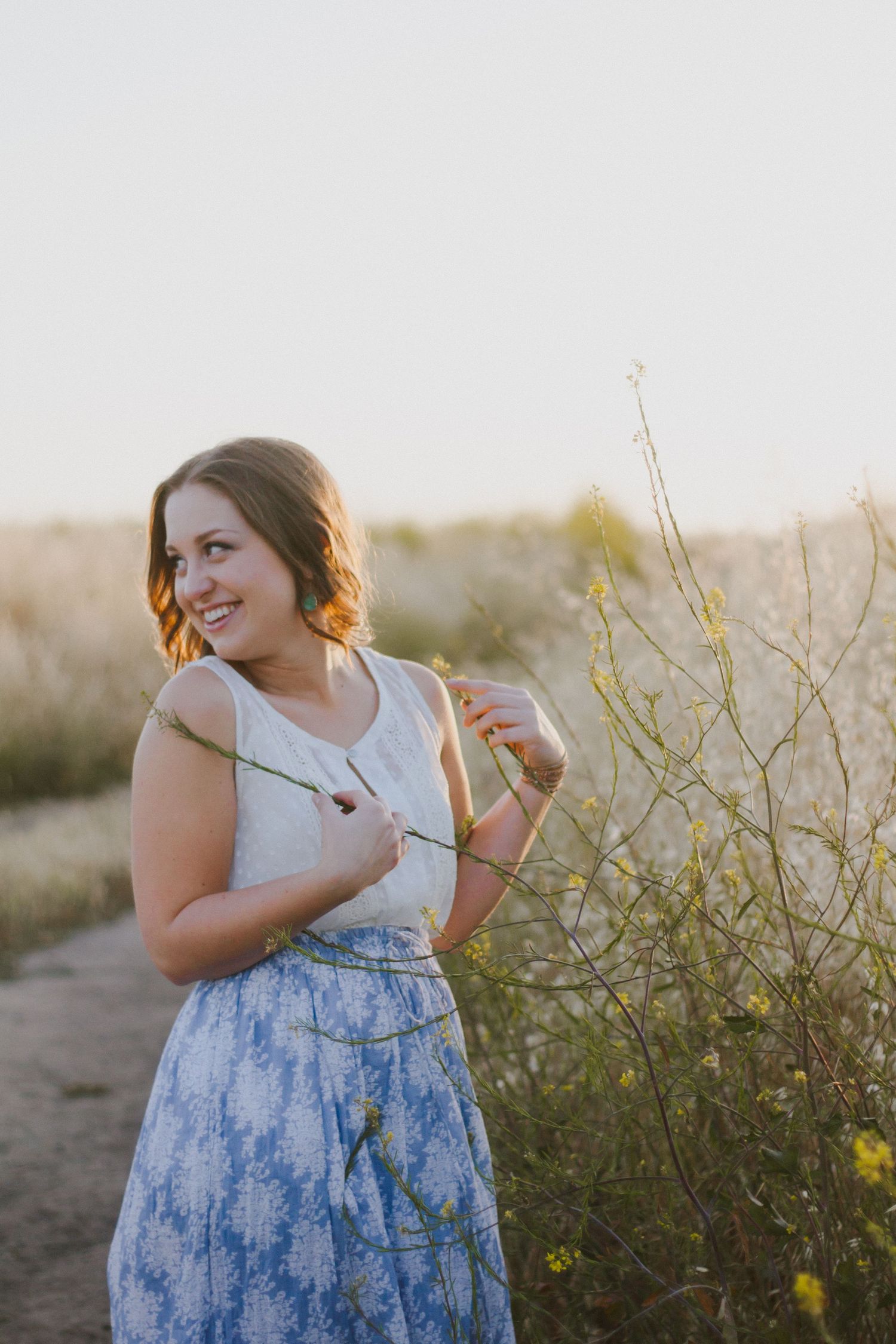 Smiling young woman in white and blue dress holding wildflowers in sunny field.
