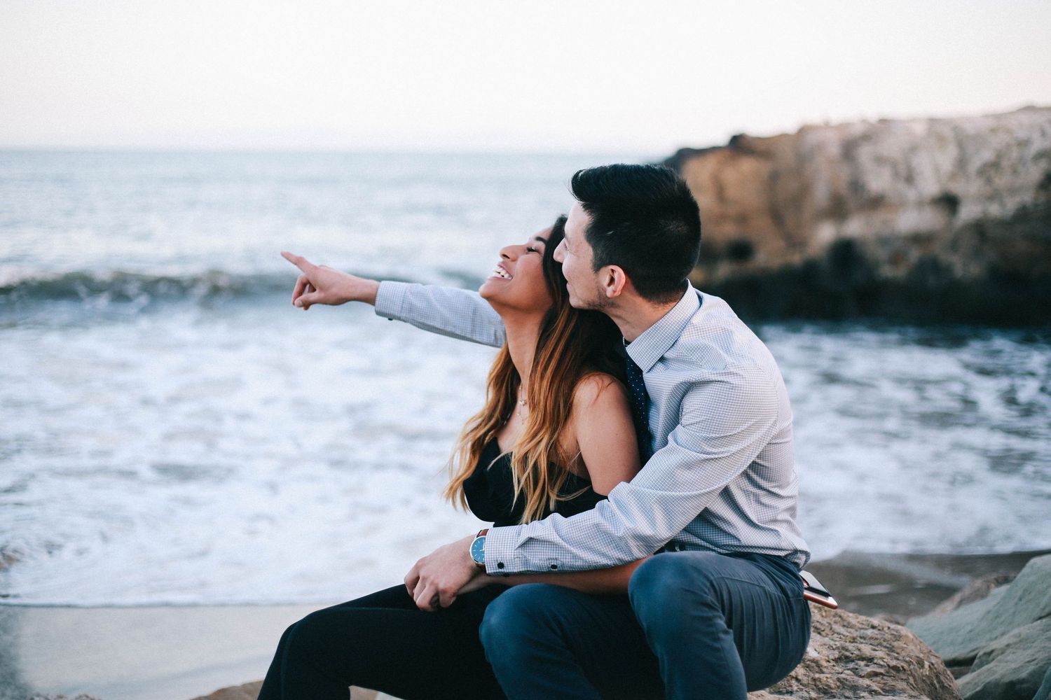 Couple sitting on coastal rocks at dusk, woman pointing toward the ocean waves in the distance.