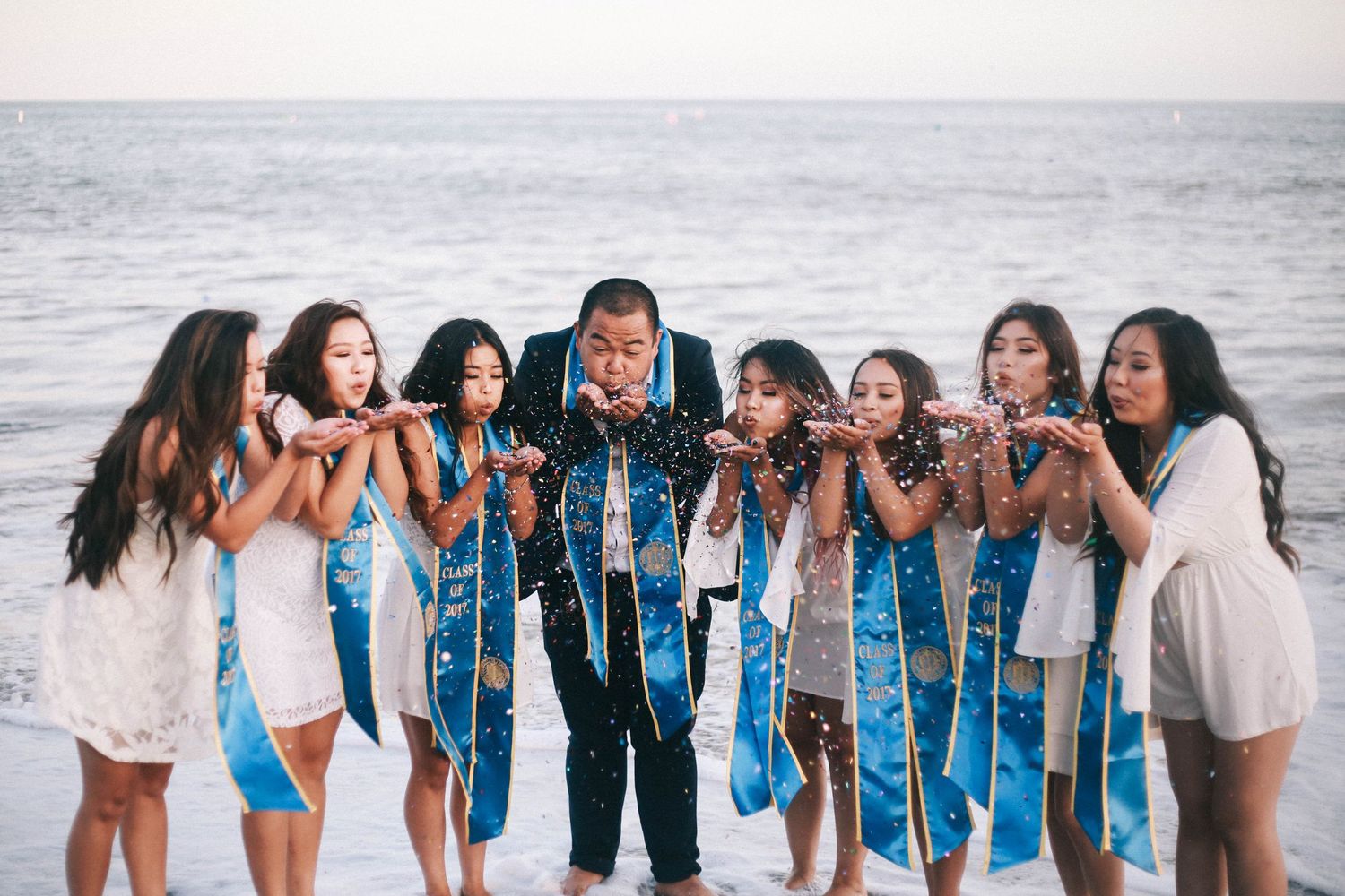 Graduate in cap and gown surrounded by smiling women in blue sashes blowing confetti at the beach.