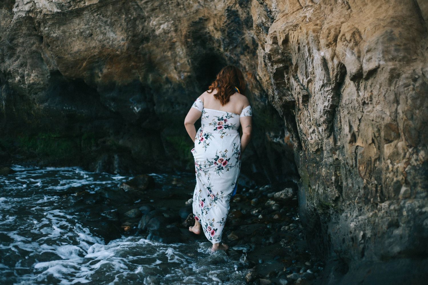 Woman in floral off-shoulder maxi dress standing against a rocky cliff by the ocean.