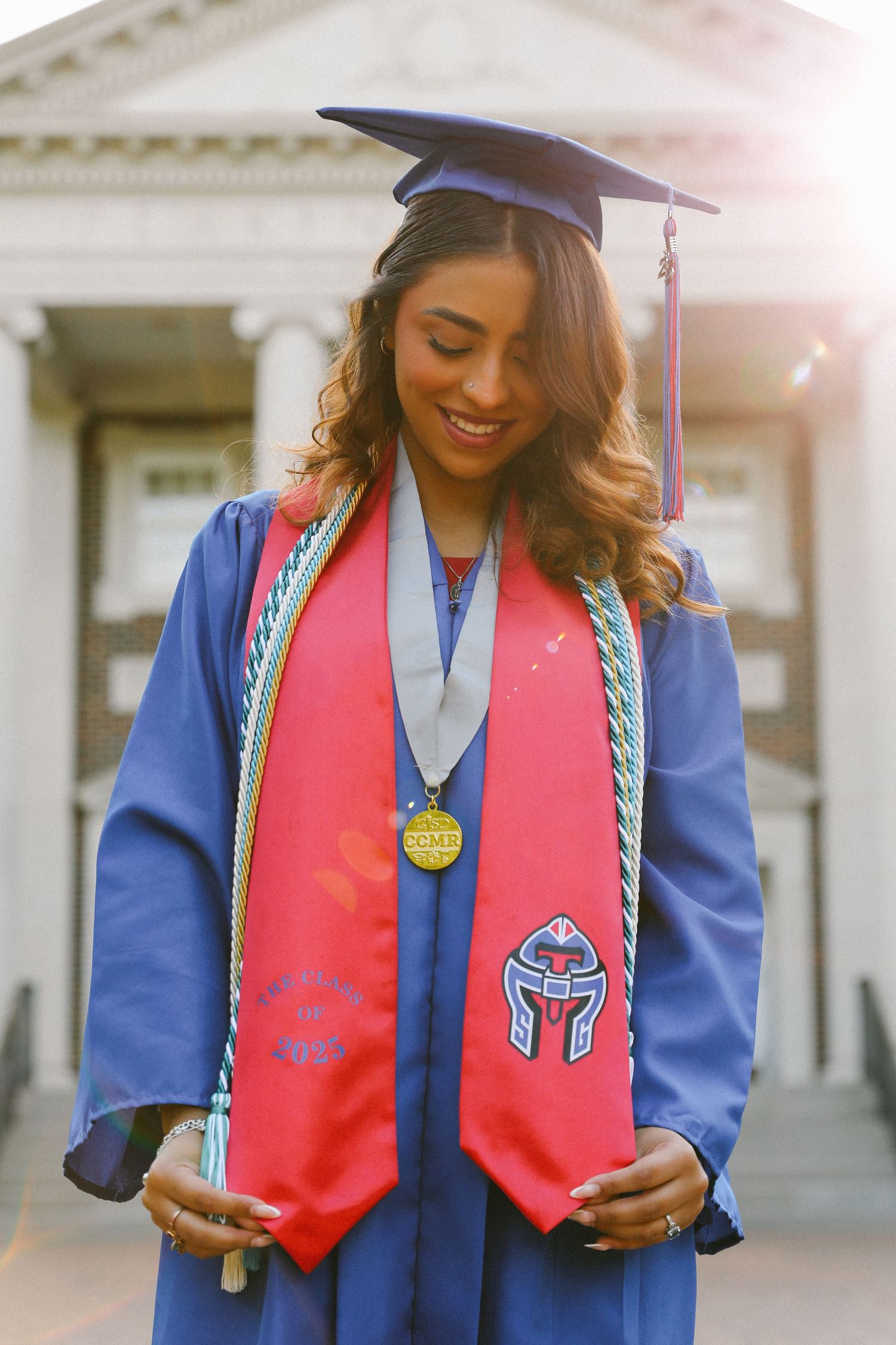Graduate in blue cap and gown with red stole and medal posing in front of a columned building.