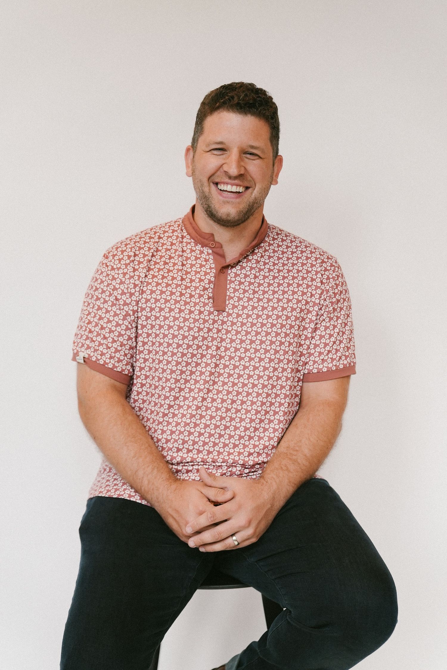 Smiling man in patterned pink polo shirt sitting against a white studio background.