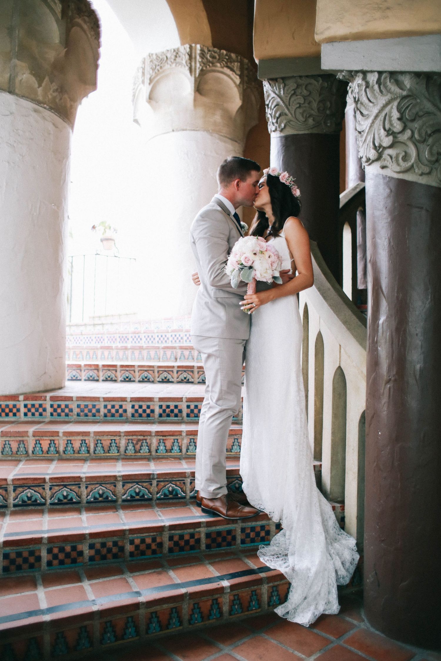 Wedding couple kissing on decorative tiled staircase with ornate stone columns in elegant venue.