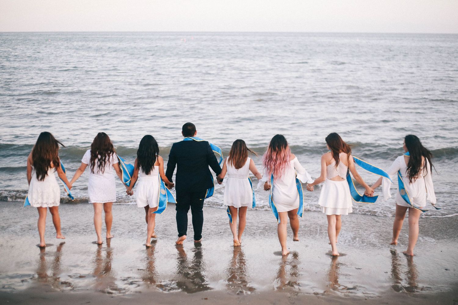 Group of friends in white dresses holding hands at beach shore during golden hour.