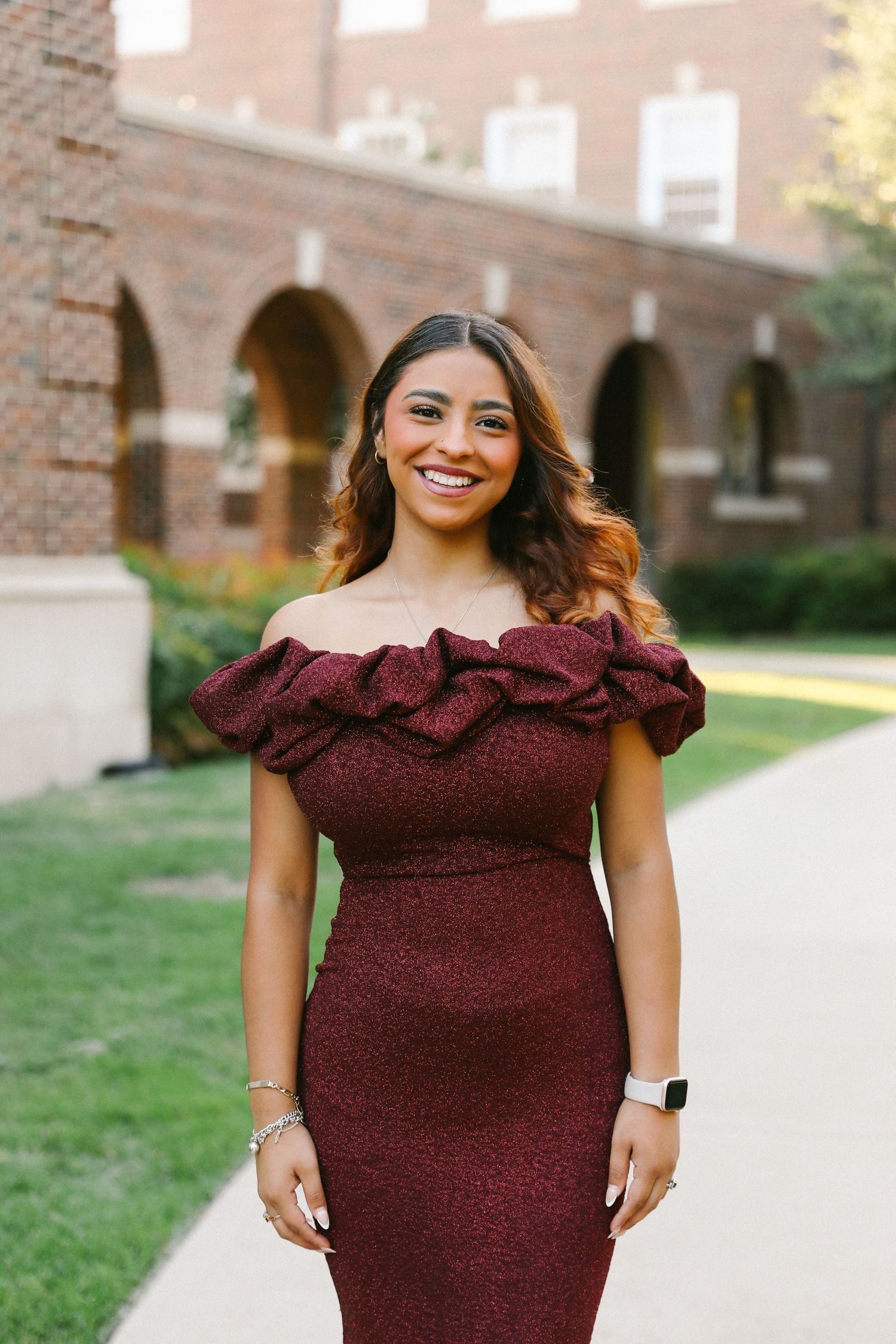 Smiling young woman in off-shoulder burgundy dress posing in front of a brick archway outdoors.