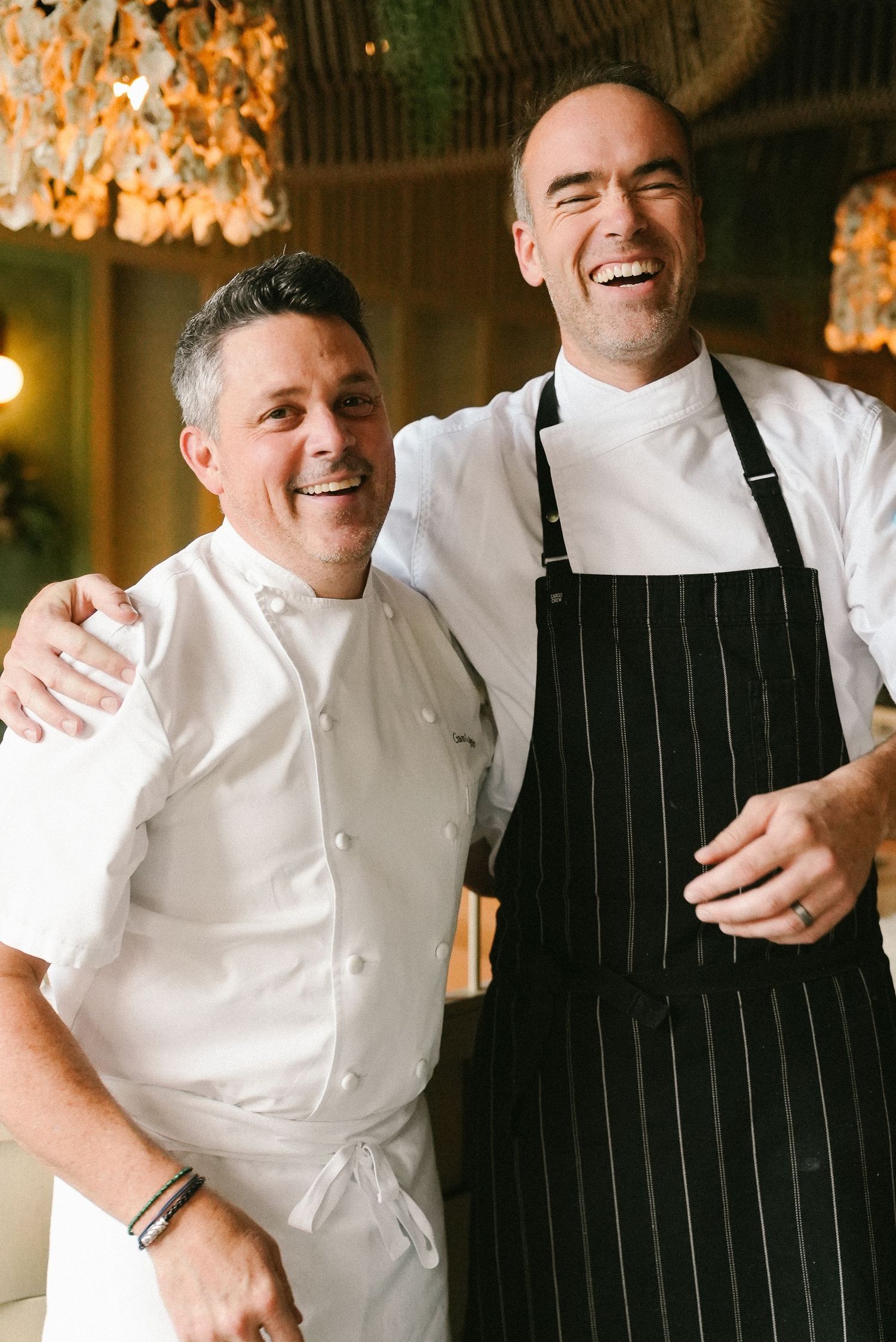 Two smiling chefs in white jackets and aprons pose together in an elegant restaurant setting.