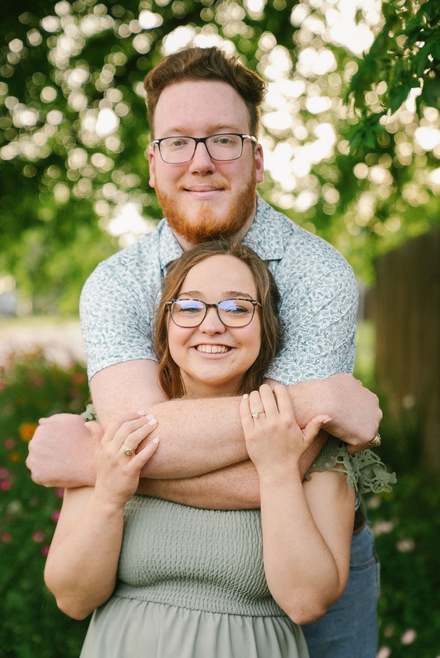 A happy couple embraces outdoors, man with red beard hugging woman wearing glasses and sage green dress.