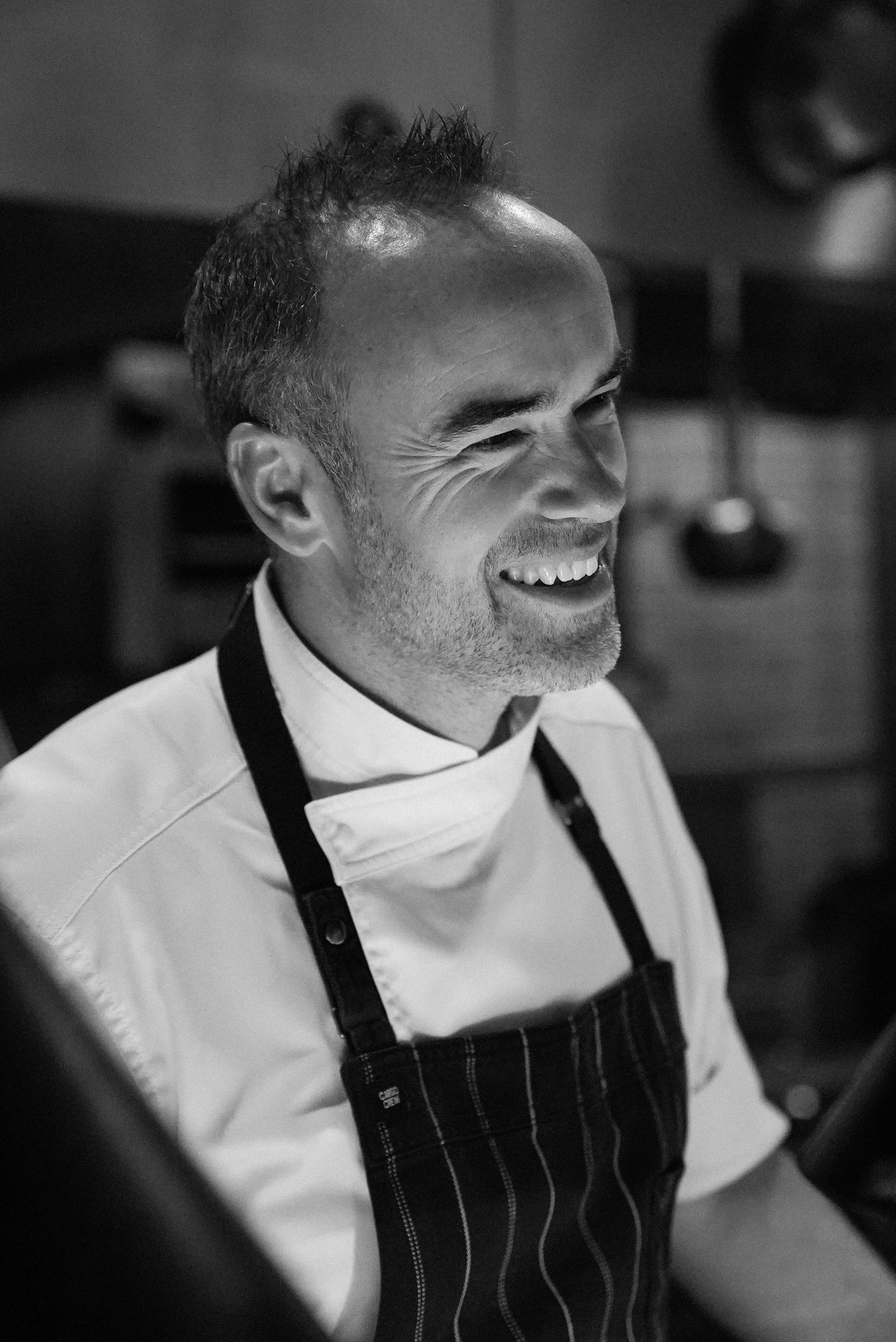 Smiling male chef in white uniform and striped apron photographed in black and white in a professional kitchen.