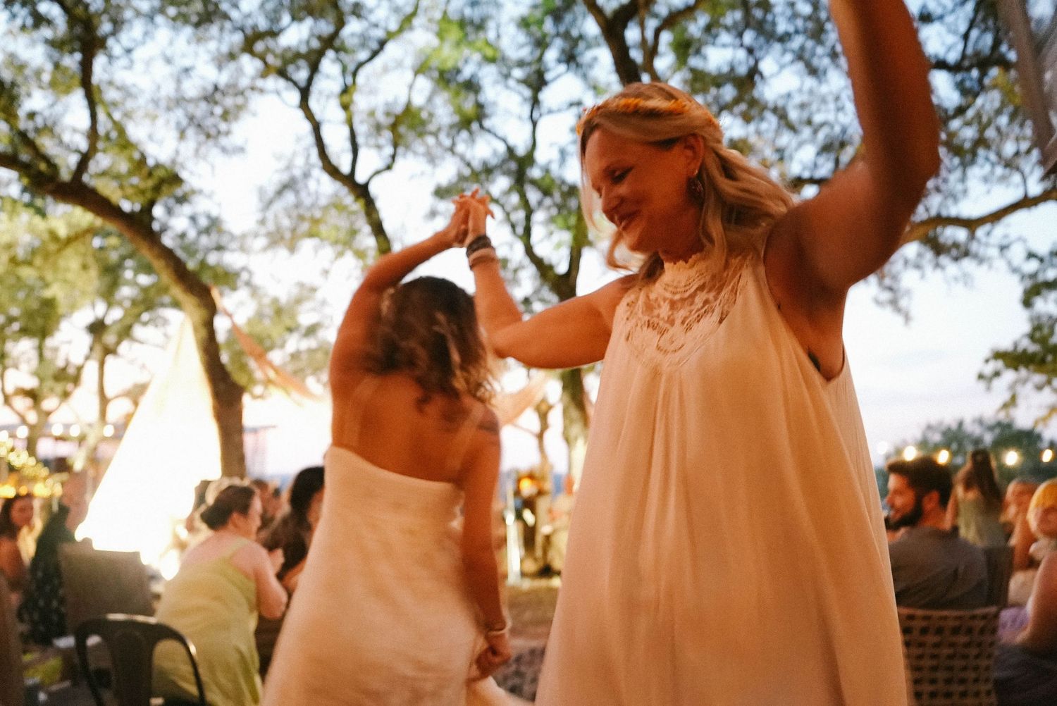 Two brides in white dresses dance together joyfully at an outdoor wedding reception under oak trees.