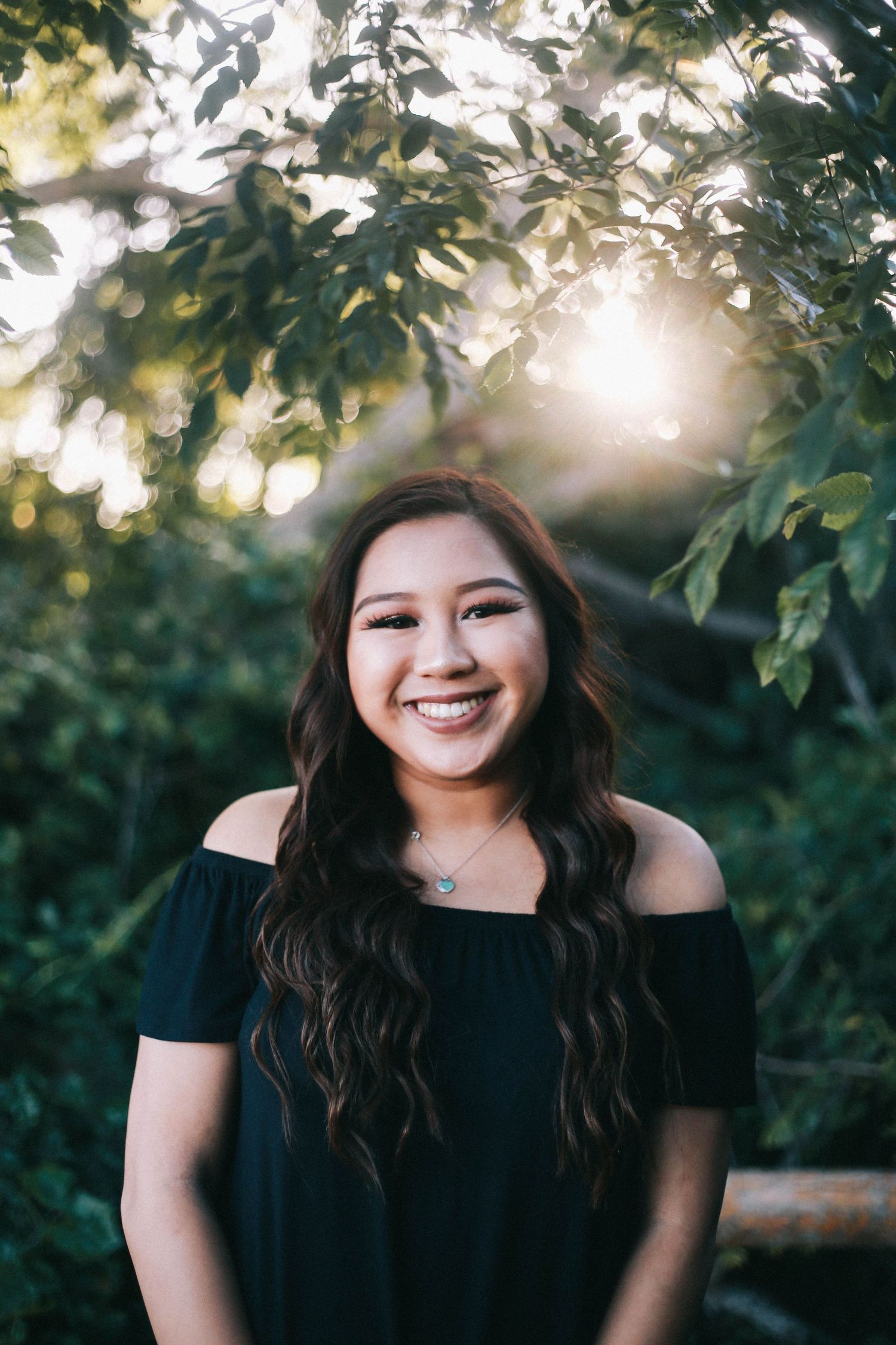 Smiling young woman in black off-shoulder top posing outdoors in sunlit garden.