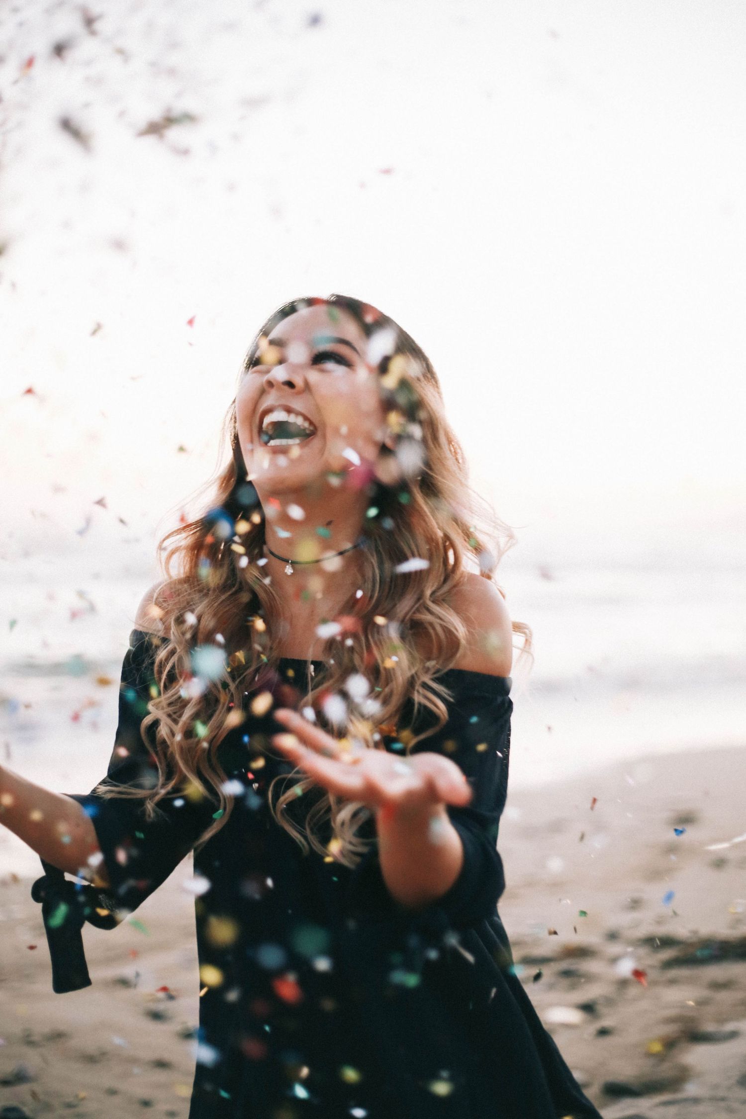 Joyful woman laughing as colorful confetti falls around her on a beach.