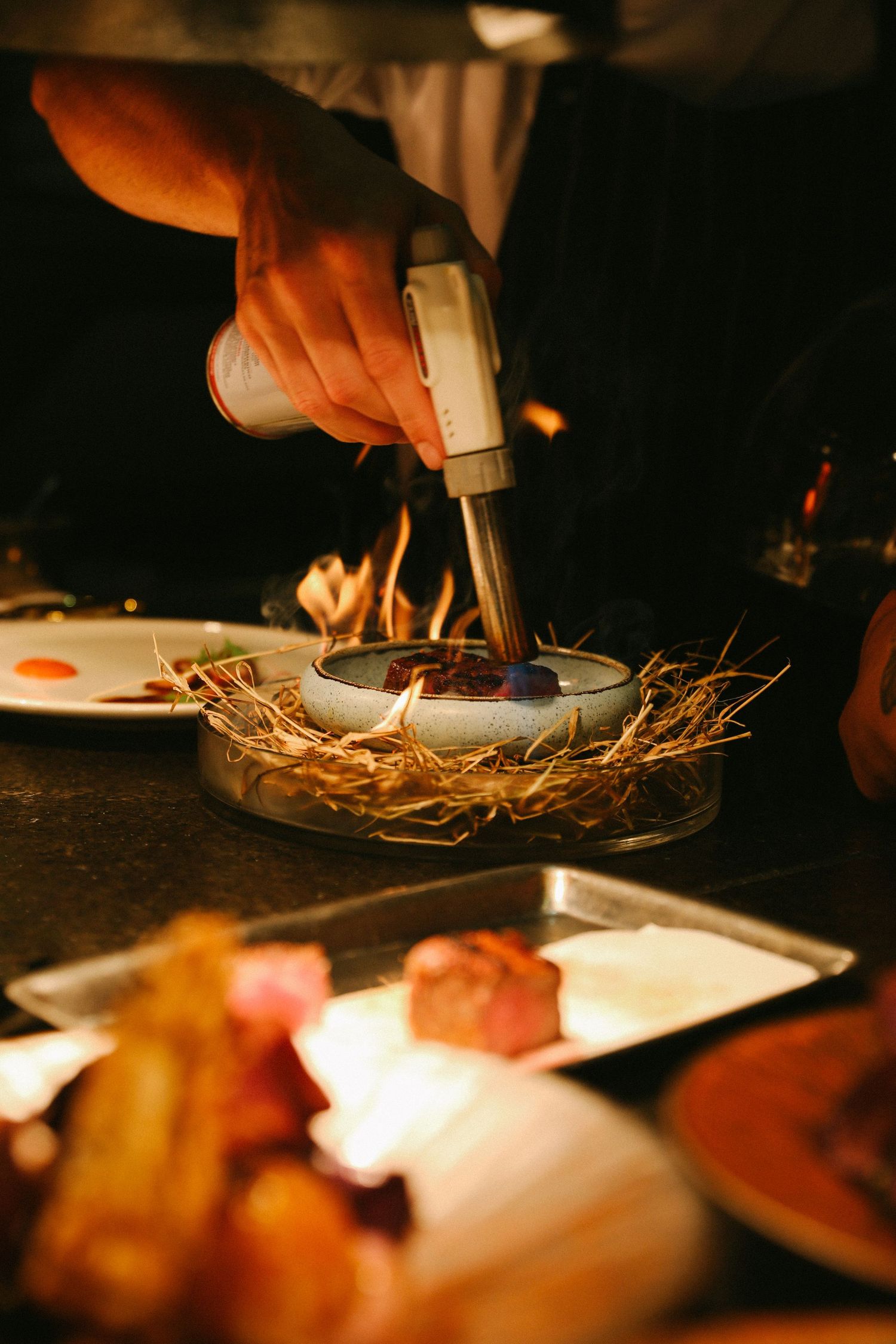 Chef using blowtorch to flame finish a gourmet dish nestled in hay in restaurant kitchen.