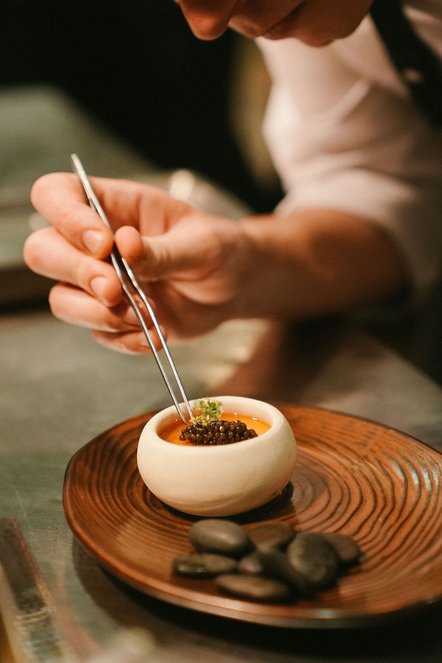 Chef using tweezers to plate a gourmet dish in a white bowl on a wooden plate with stones.