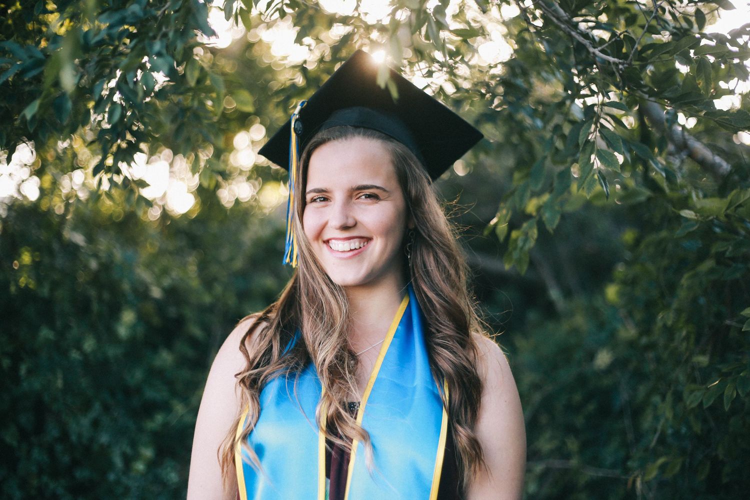 Happy young woman in graduation cap and blue and gold stole smiling under green trees.