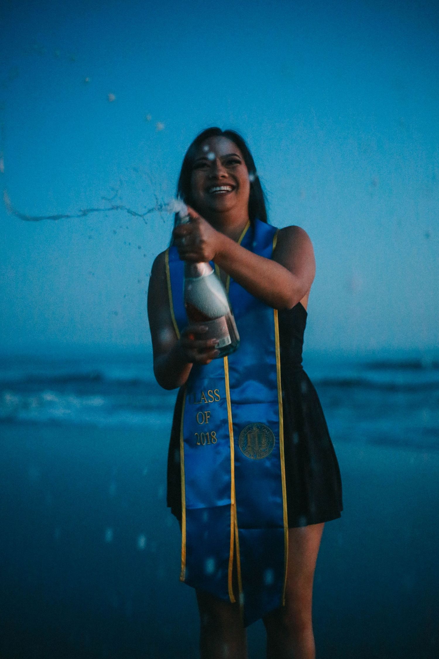 Joyful graduate in UCSB blue stole popping a champagne bottle on the beach at dusk.