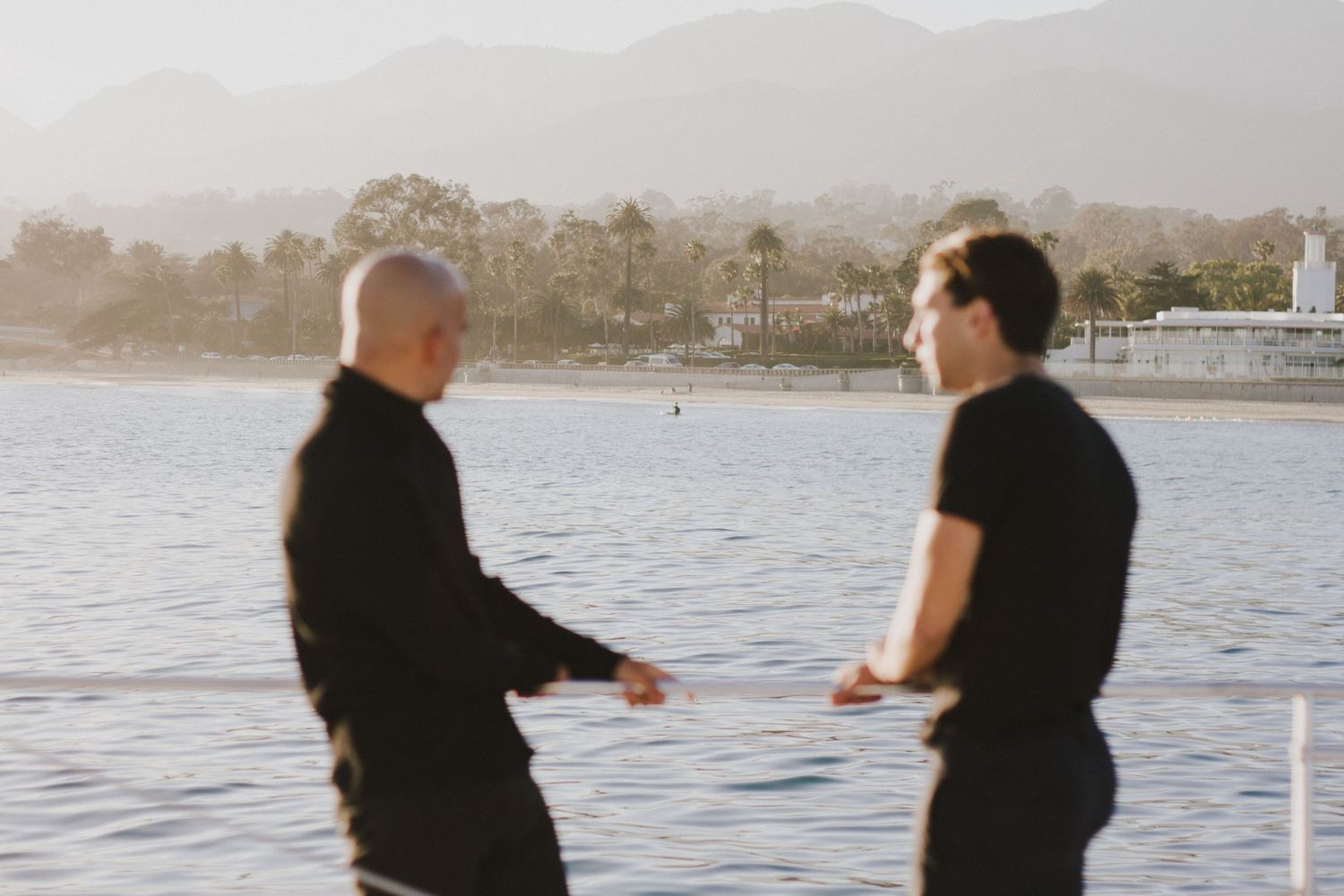 Two men in black stand facing each other near a calm waterfront with mountains in the background.