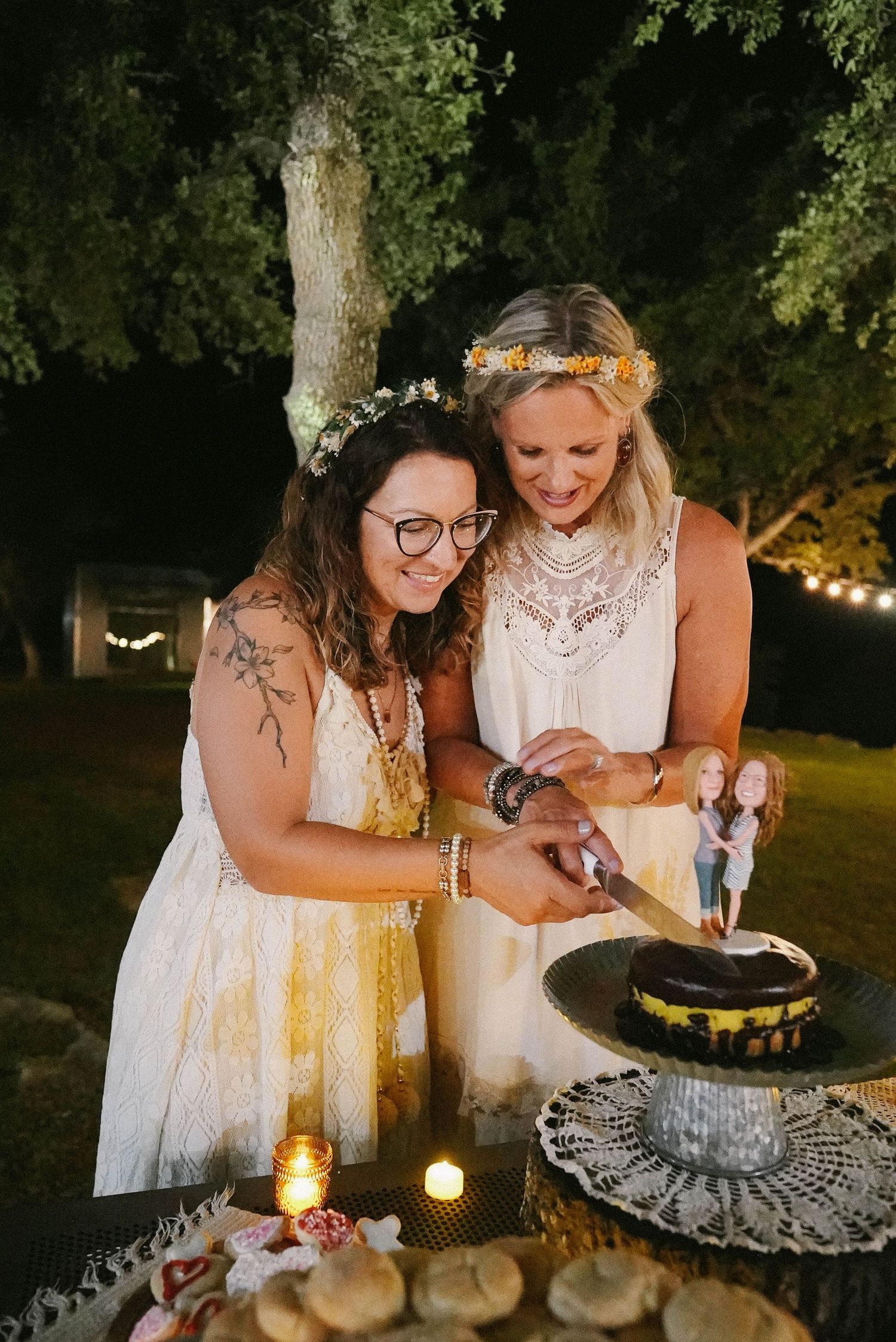 Two brides wearing flower crowns smile while cutting their wedding cake together at an outdoor night reception.