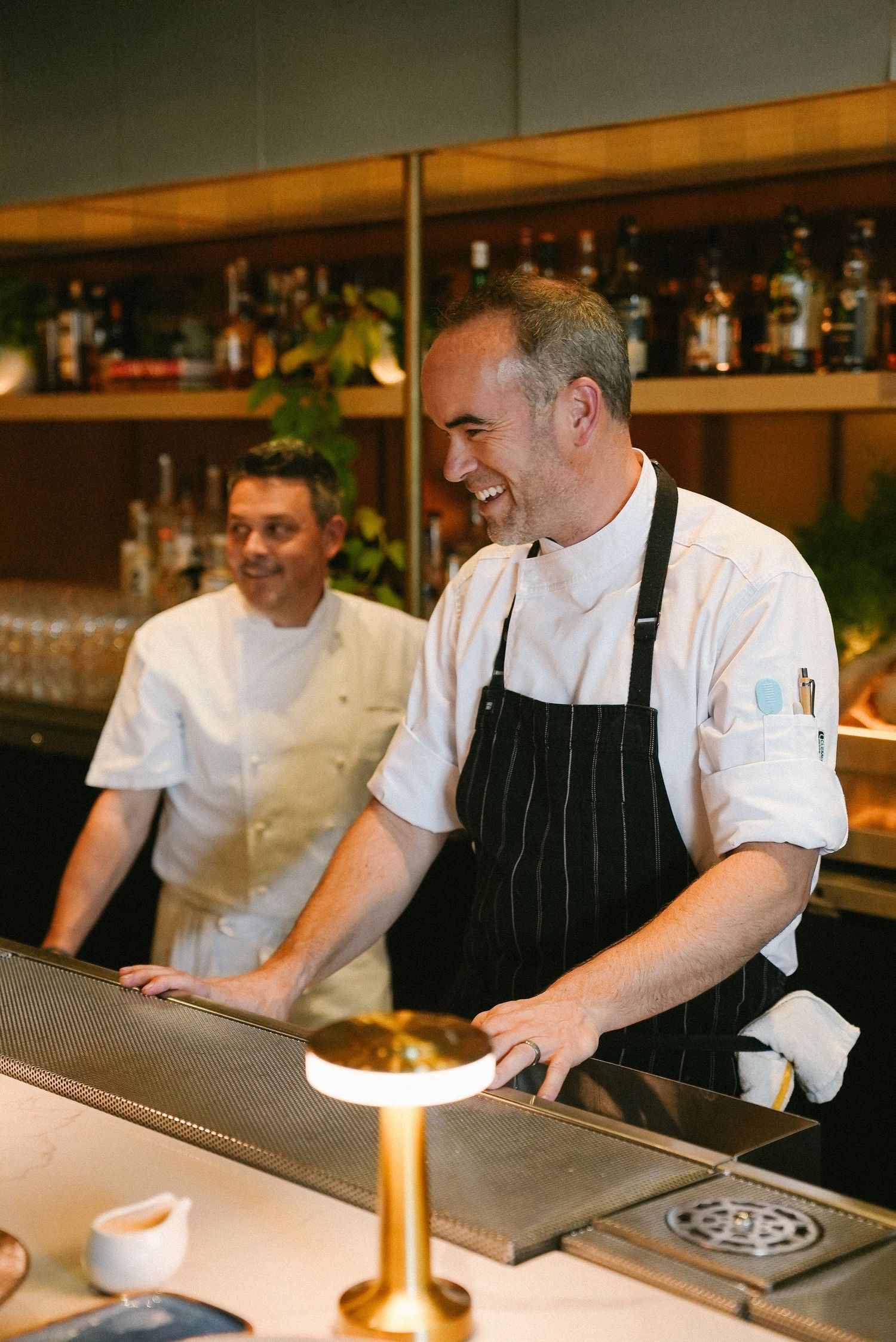 Two professional chefs in white uniforms and black aprons working behind a restaurant counter.