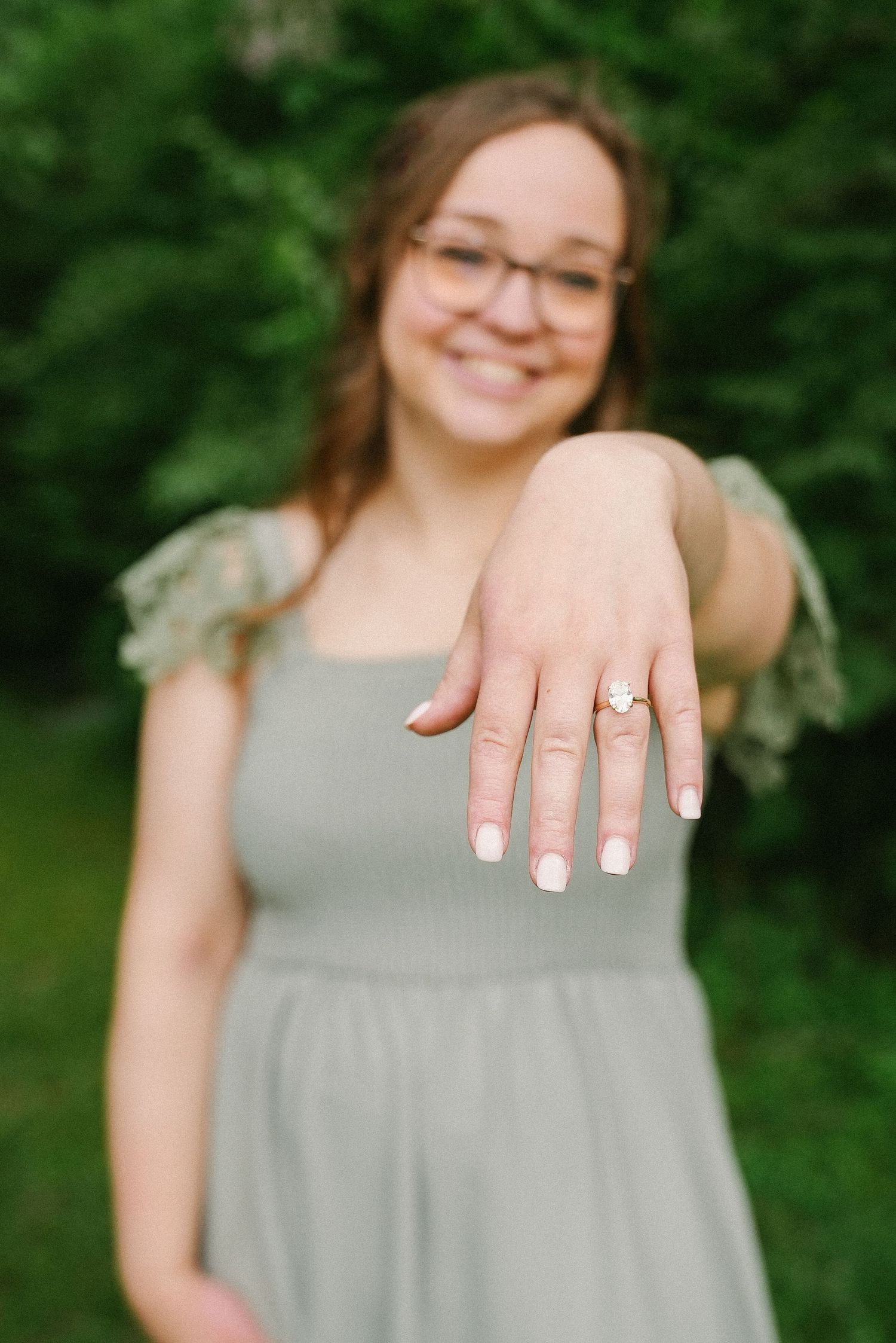 Smiling woman in sage green dress shows off engagement ring with white manicure outdoors.