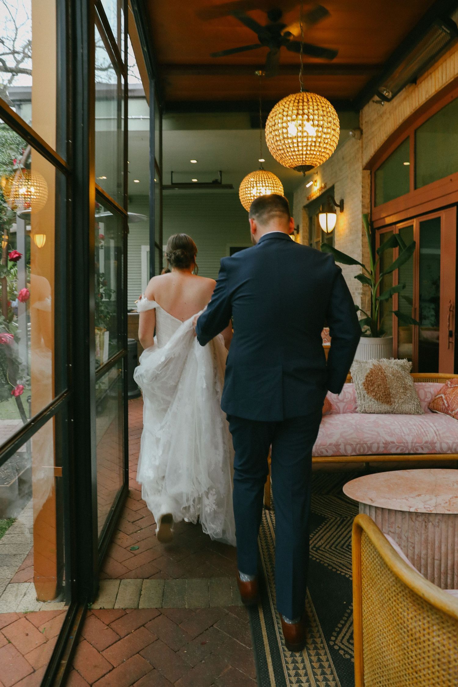 Bride and groom walking through elegant indoor-outdoor venue corridor with warm pendant lighting.