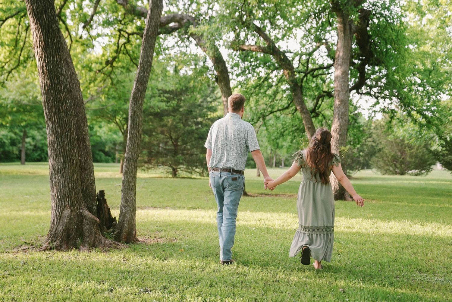 Couple holding hands walking through lush green park with large trees on sunny day.