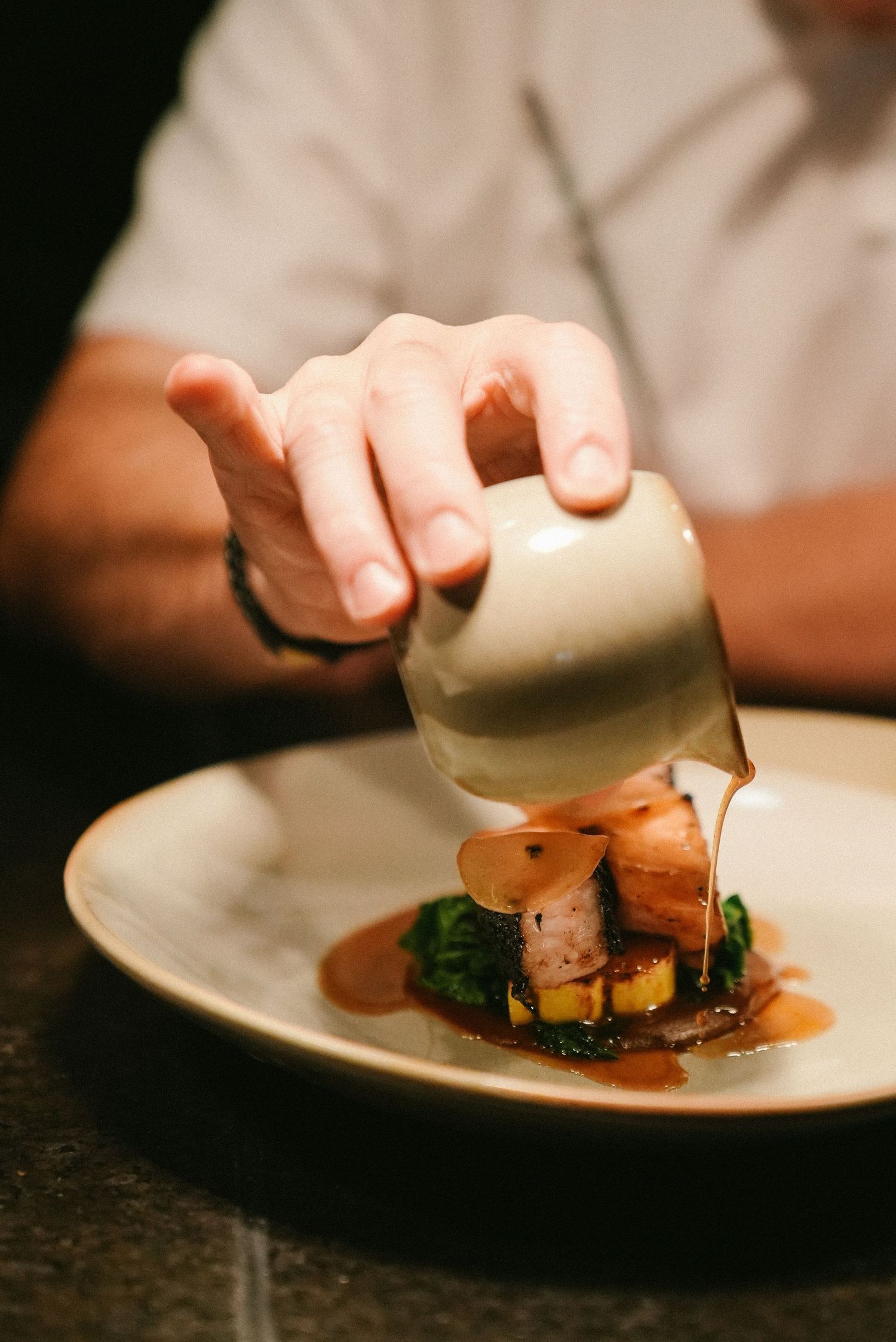 Chef pouring rich brown sauce over a gourmet fish dish with greens and mango on a ceramic plate.