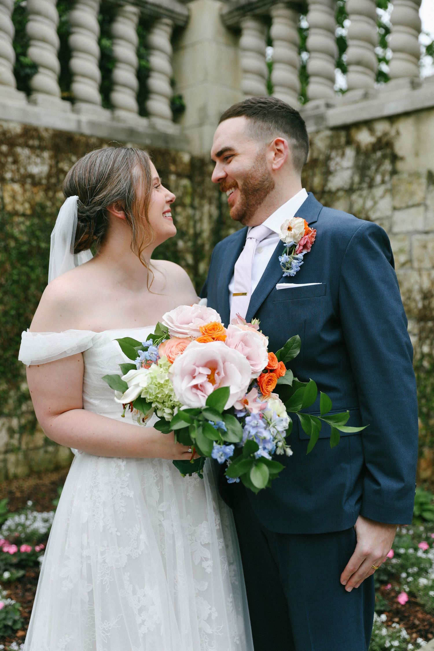 Smiling bride and groom share a joyful moment outdoors, bride holding a vibrant bouquet of pink and orange flowers.