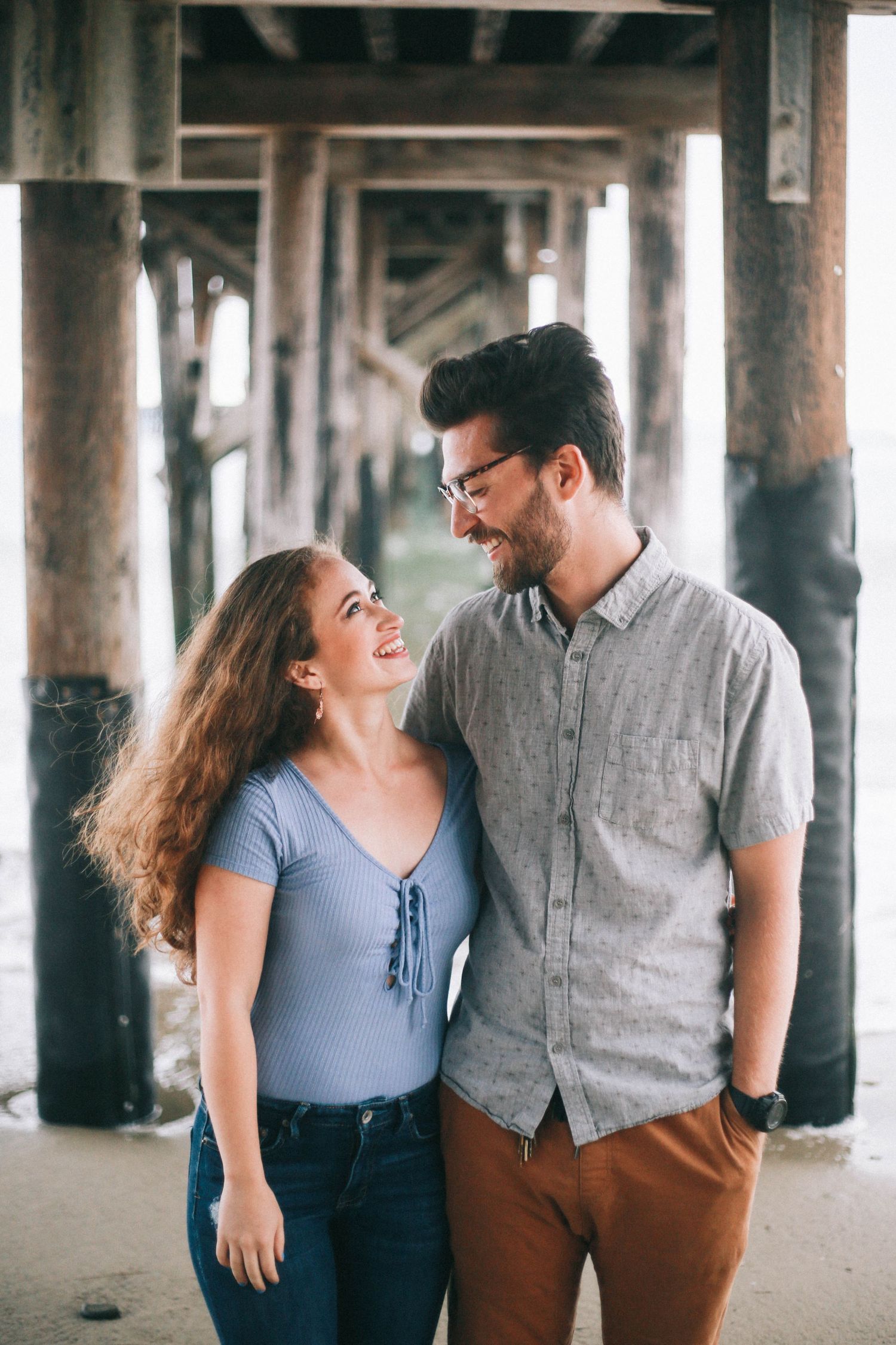 Happy couple laughs together under a wooden pier, woman in blue top and man in gray shirt.