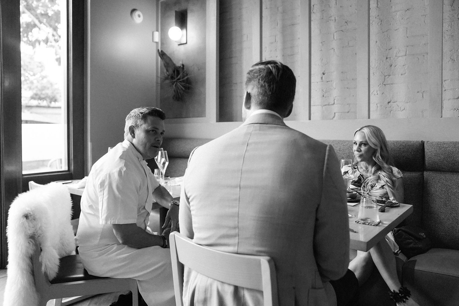 Chef in white jacket engages in conversation with dining guests in a sophisticated restaurant booth.