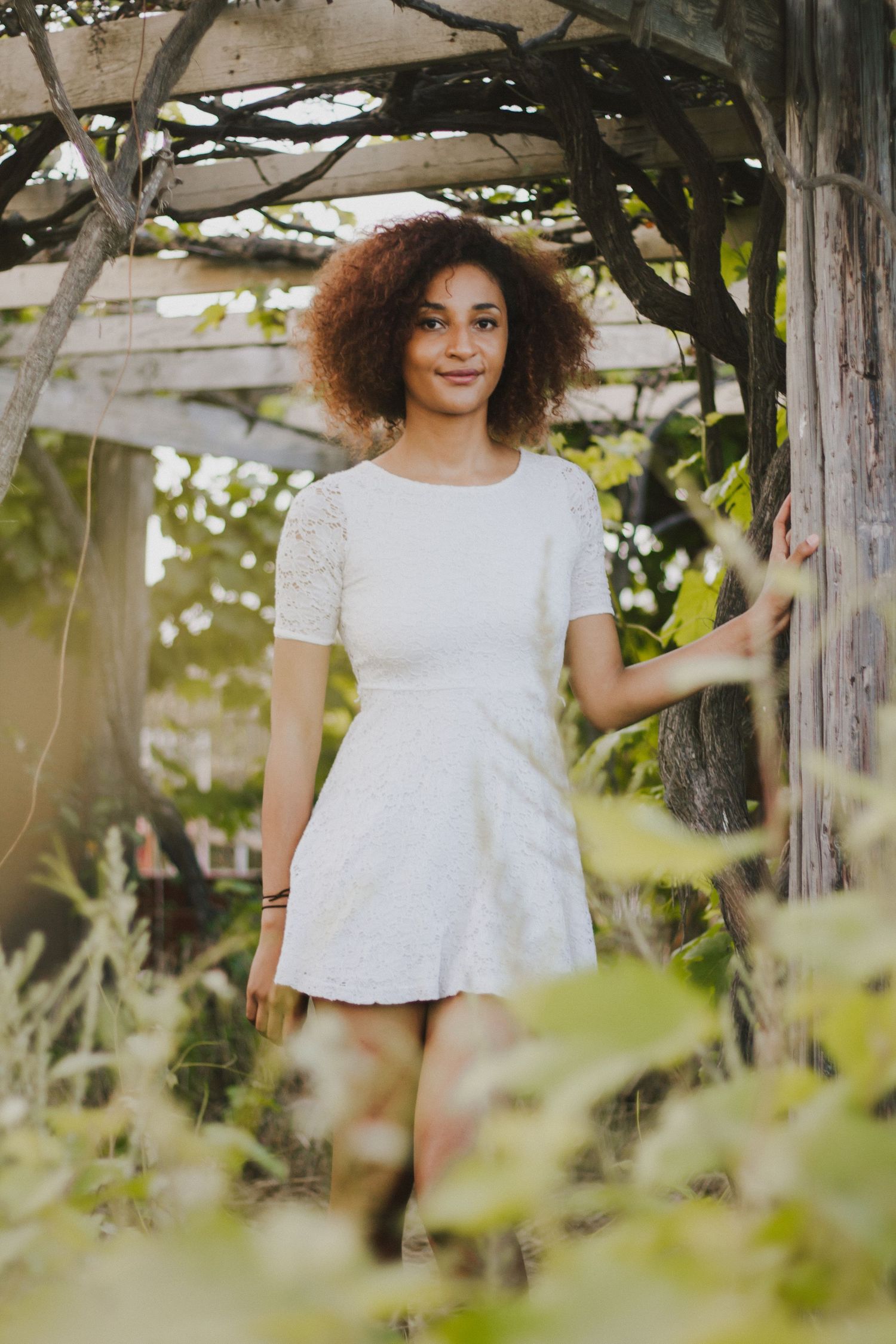 Young woman with curly hair in white lace dress stands in a lush garden pergola.