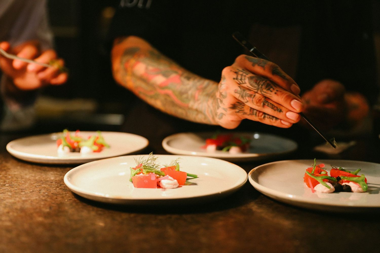 Tattooed chef plating fresh tuna tartare with tweezers on white plates in restaurant kitchen.