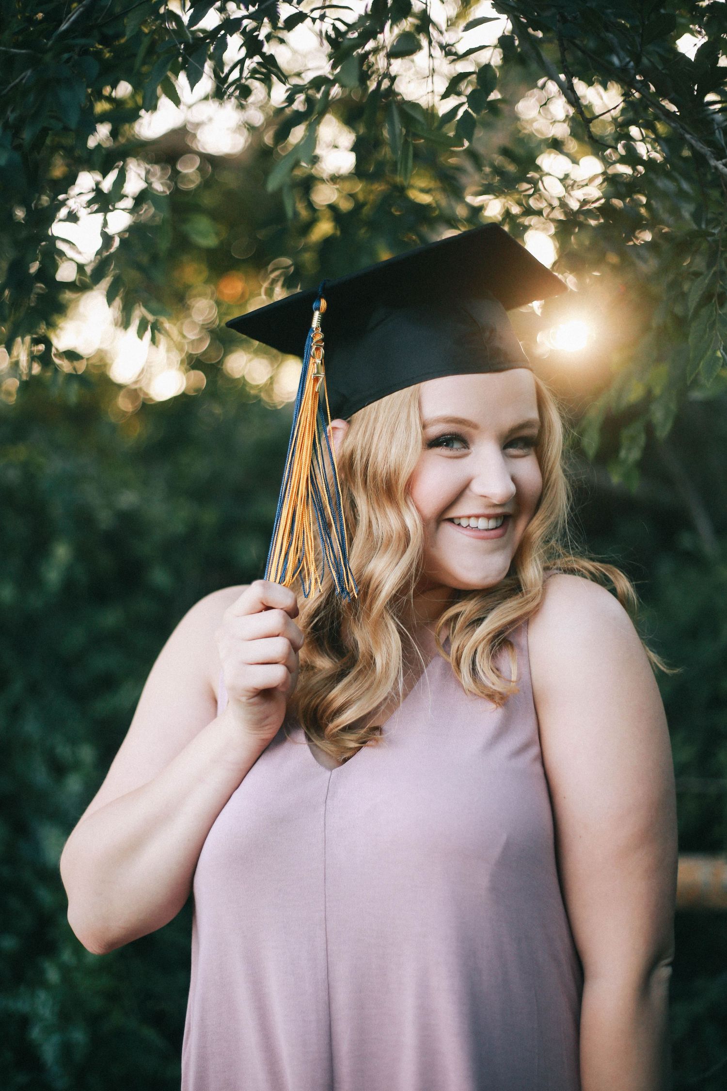 Smiling blonde woman in graduation cap holding tassel among green trees with golden bokeh light.
