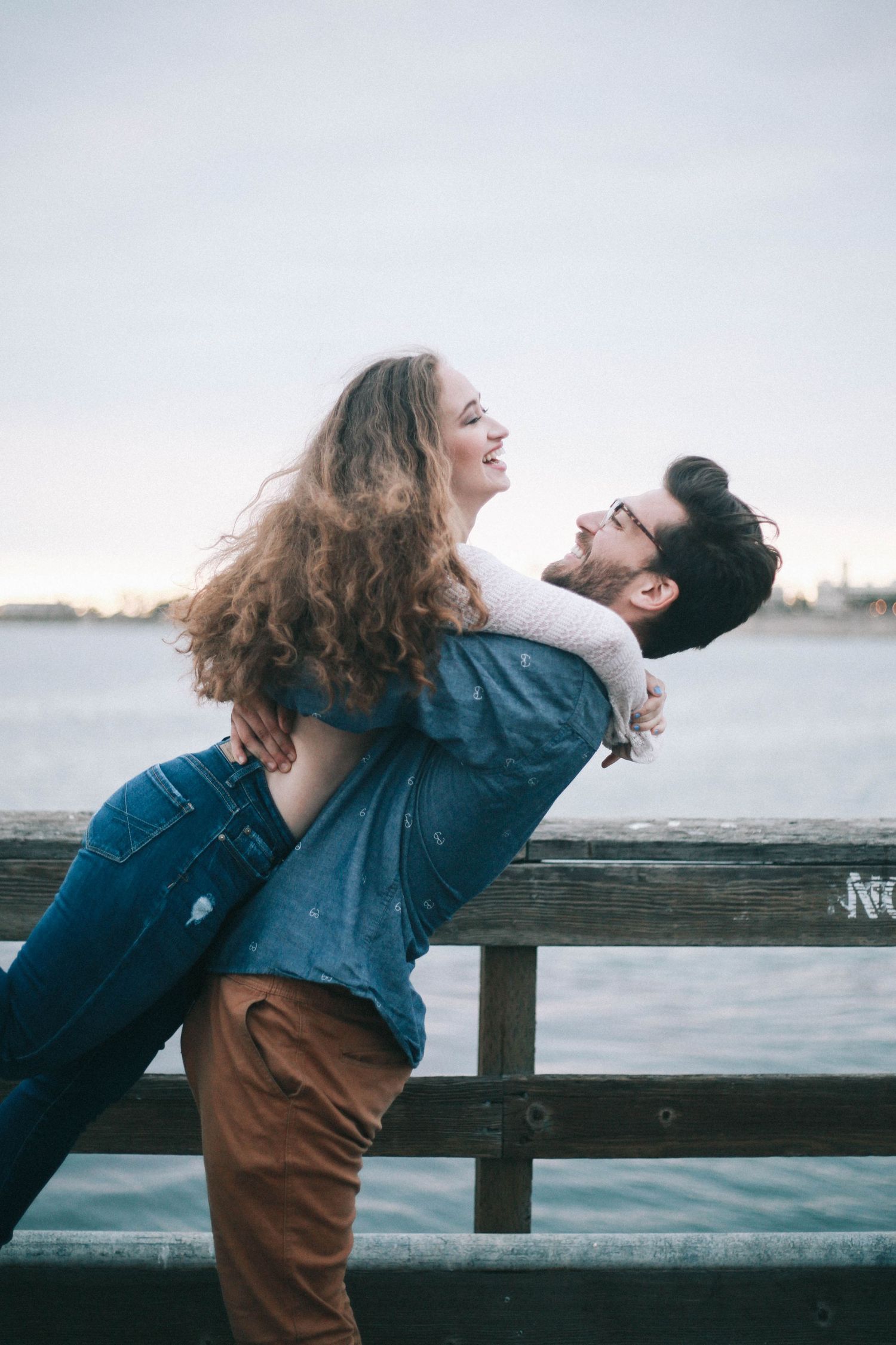 Joyful couple playfully embracing by a wooden railing with ocean in the background.