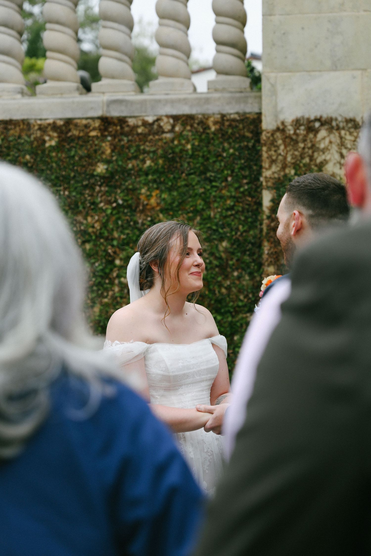 Bride smiling at groom during outdoor wedding ceremony with stone balustrade and ivy backdrop.