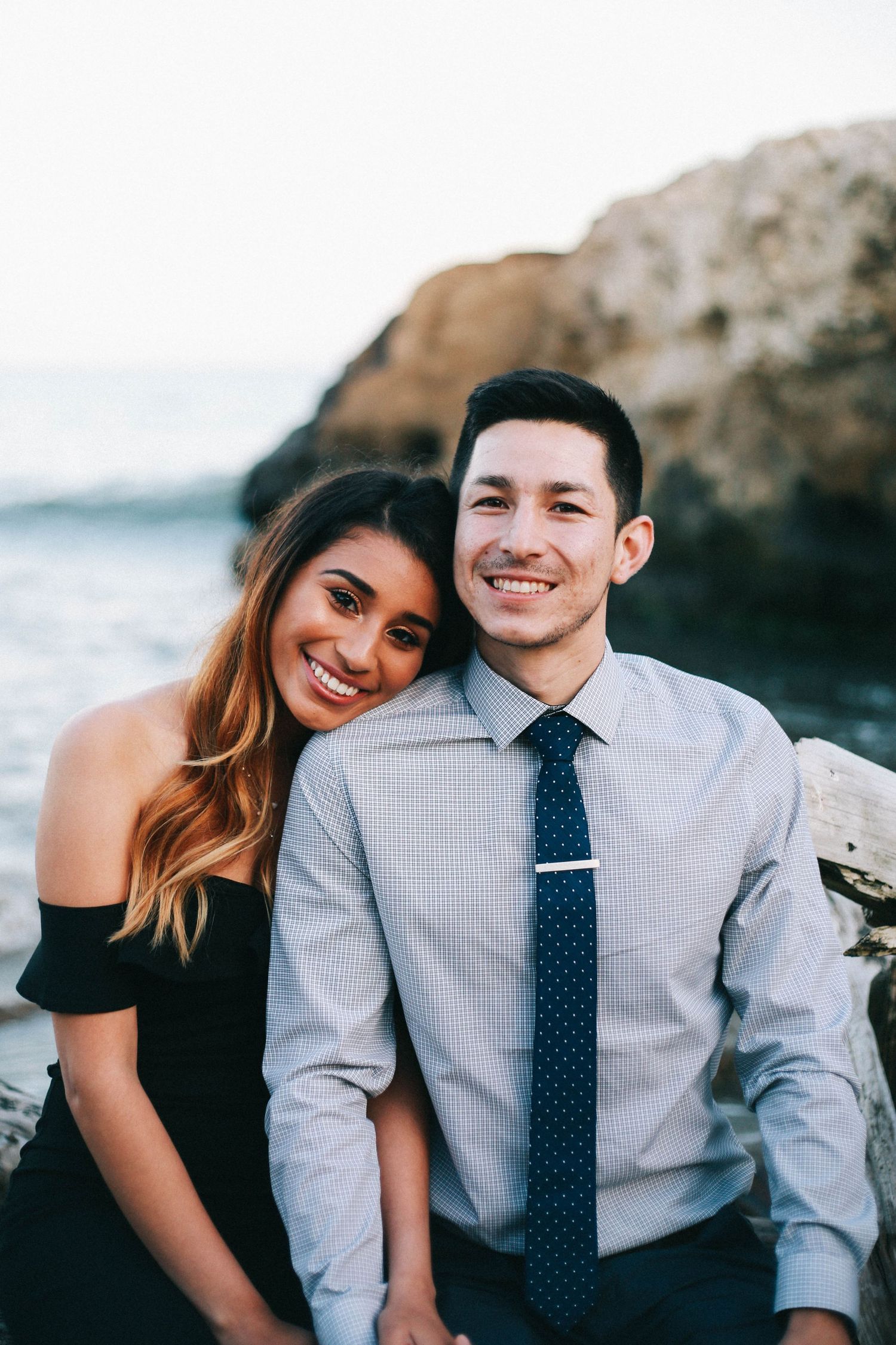 Happy couple posing together at the beach, woman in off-shoulder black top and man in gray shirt with navy tie.