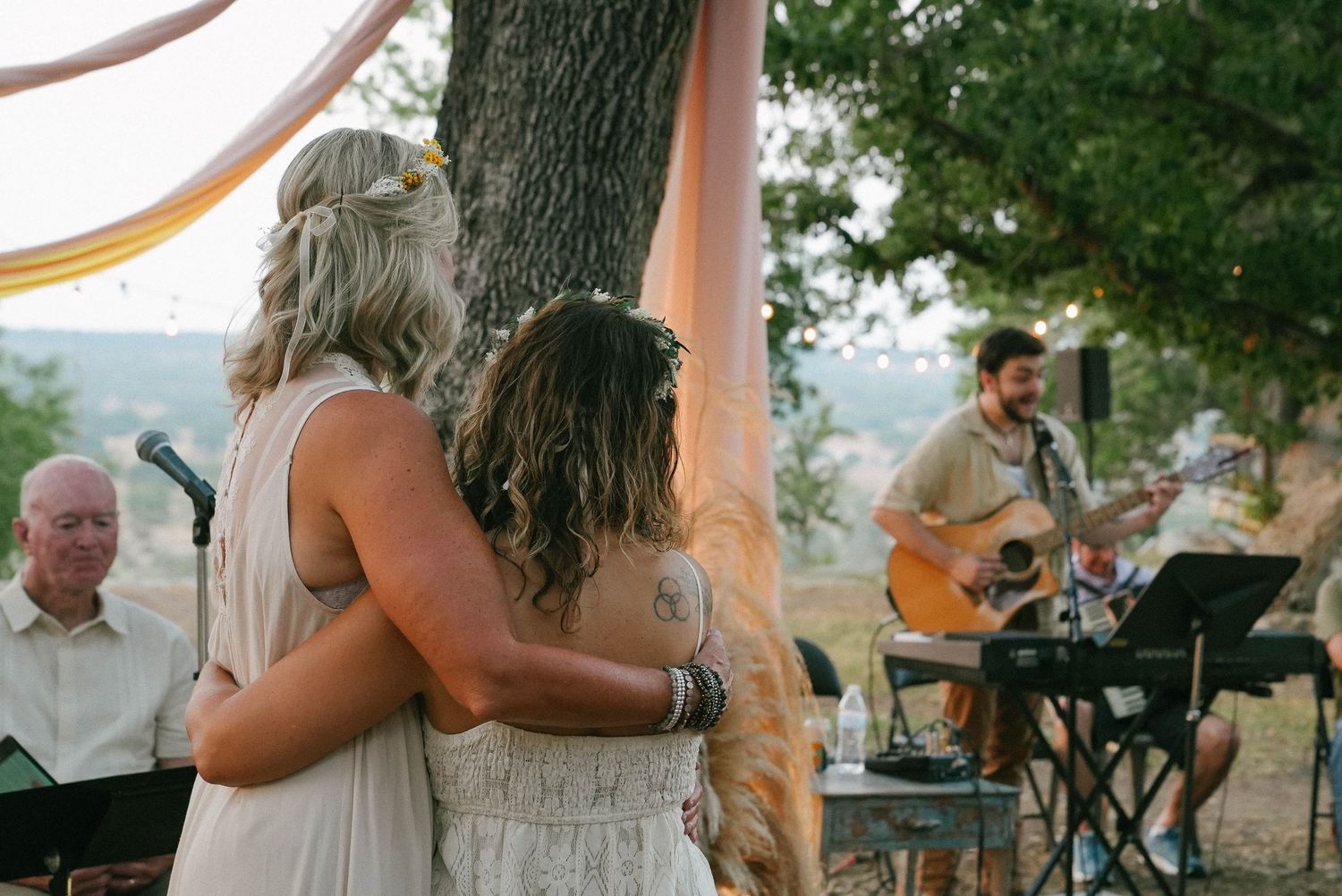 Two women embracing during outdoor bohemian wedding ceremony with live guitarist performing.