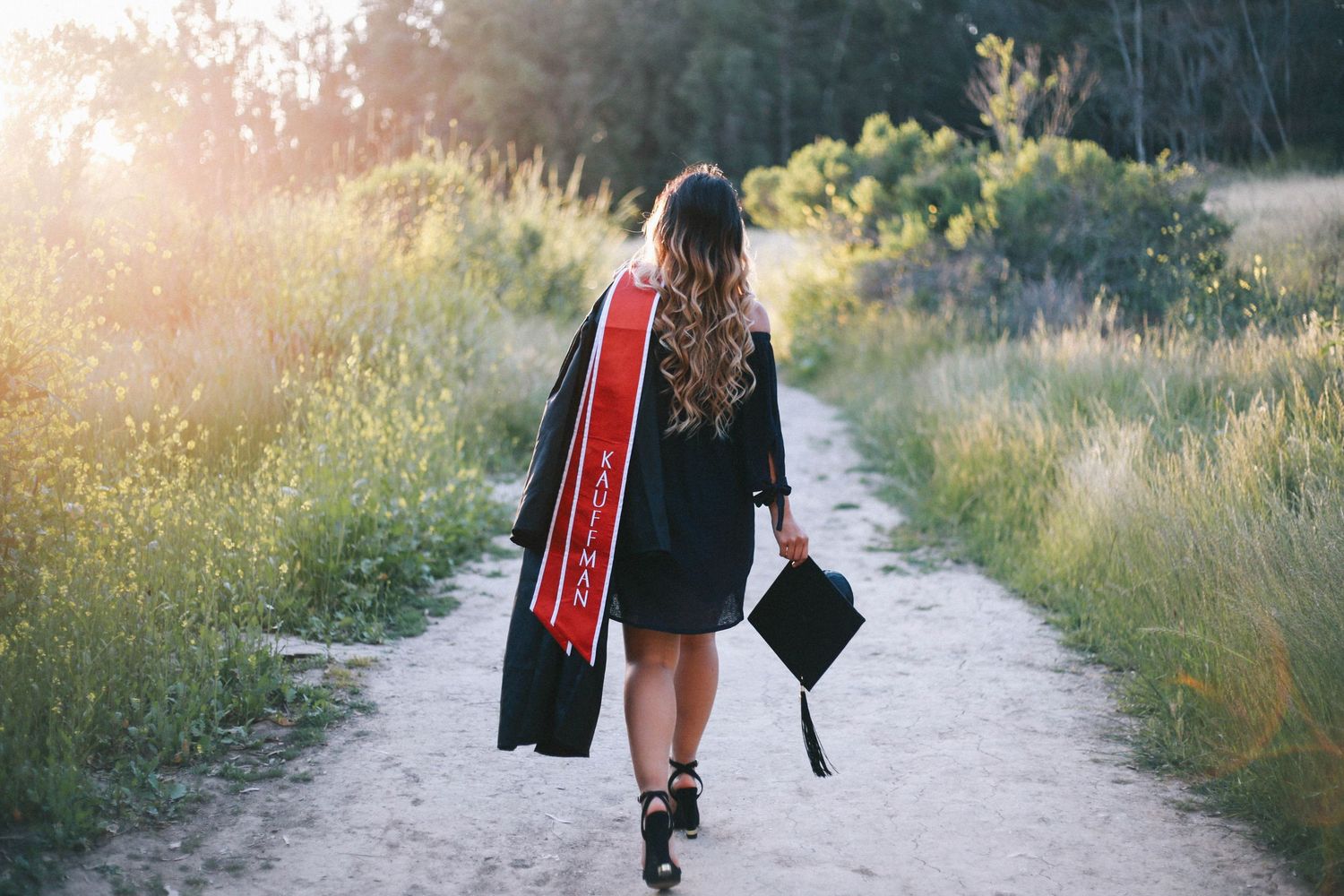 Graduate in black cap and gown with red stole walking down a sunlit nature path.