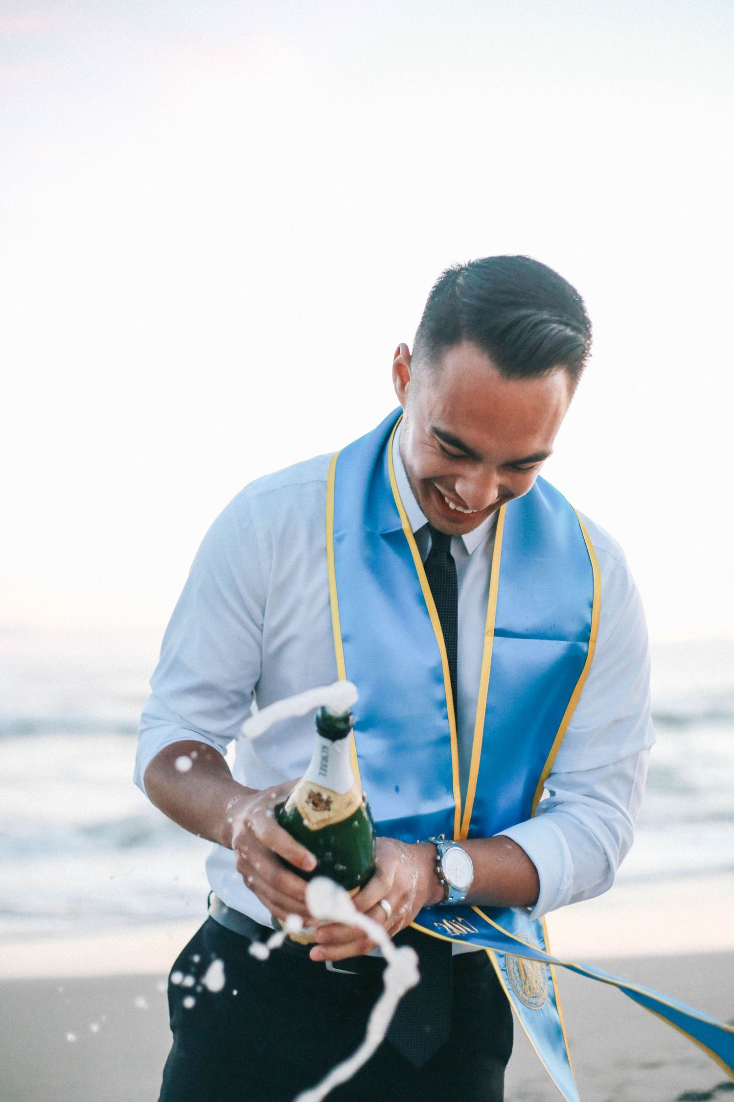 Young man in white shirt with blue graduation stole pops champagne bottle on a beach.
