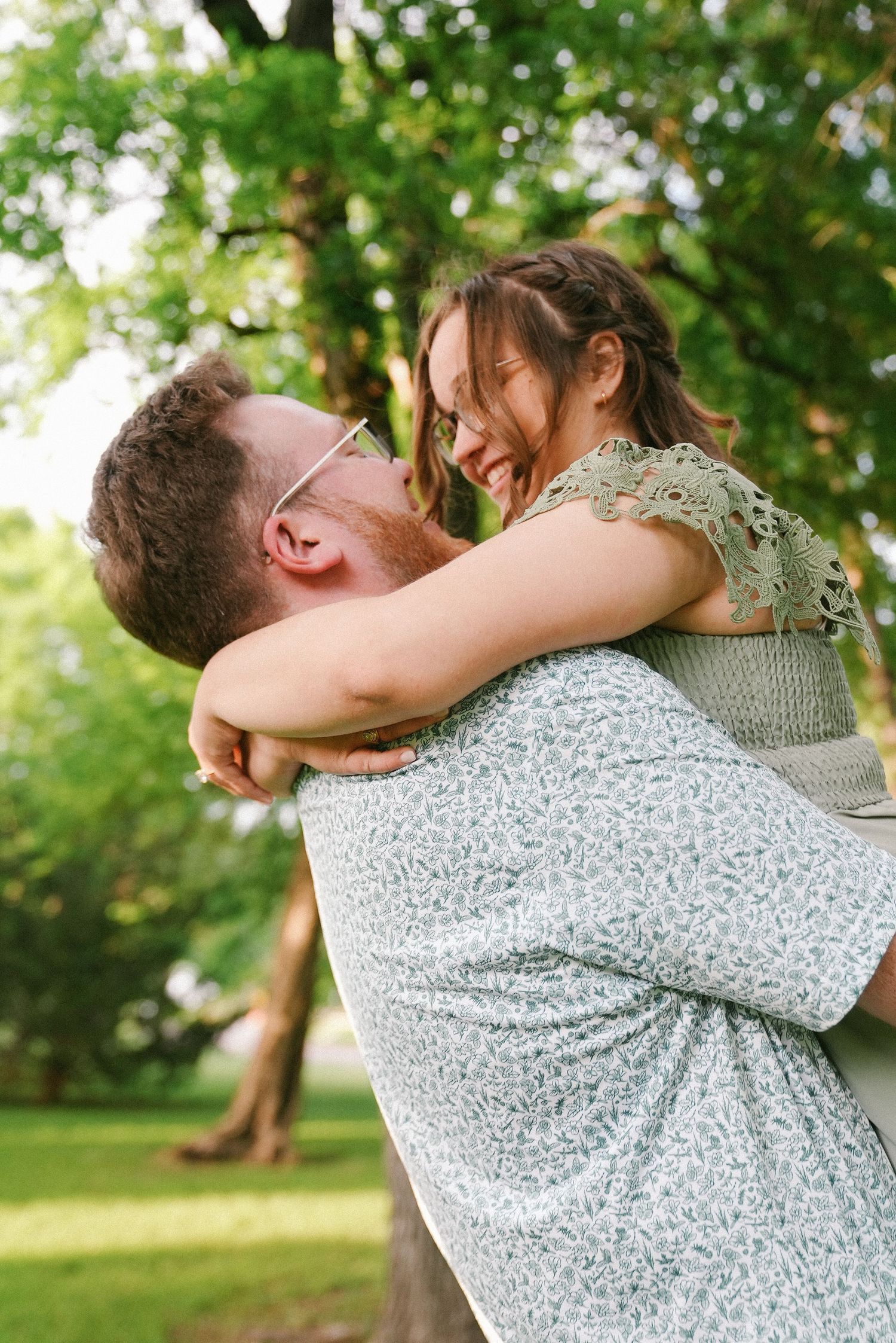 Man in floral shirt lifting and embracing laughing woman in sunny green park setting.