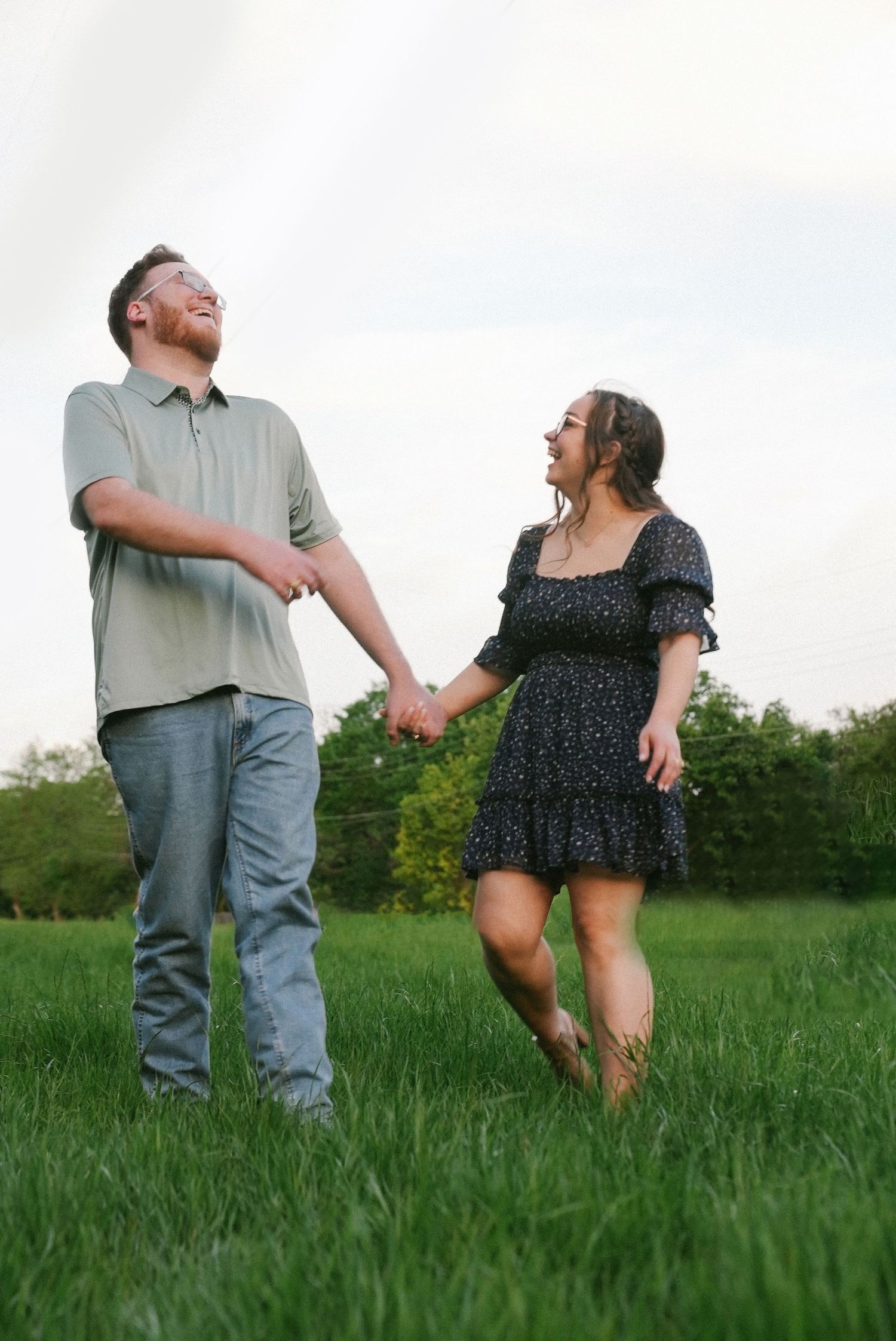 Joyful couple holds hands while walking through a lush green field at dusk.