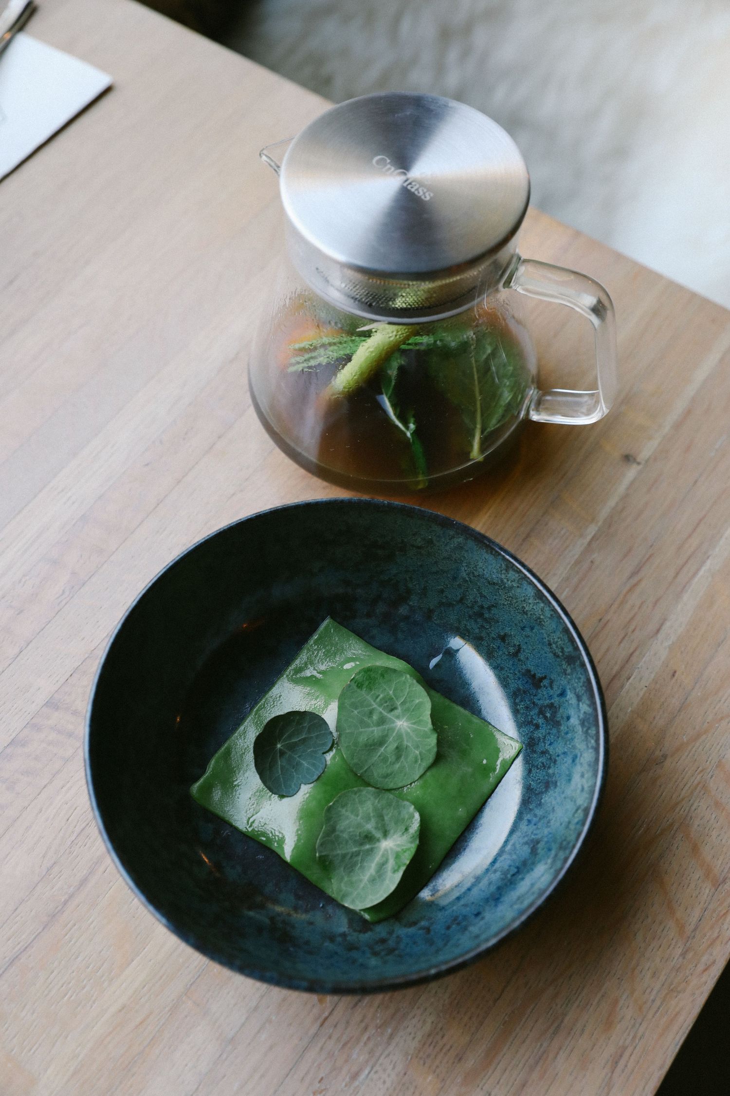 Fresh herbal tea in glass teapot with green leaf wrapped dish in ceramic bowl.