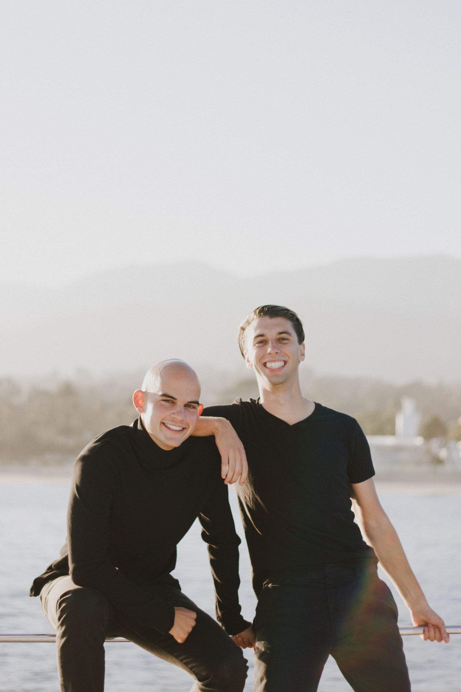 Two young men in black outfits posing together near a waterfront with mountains in background.