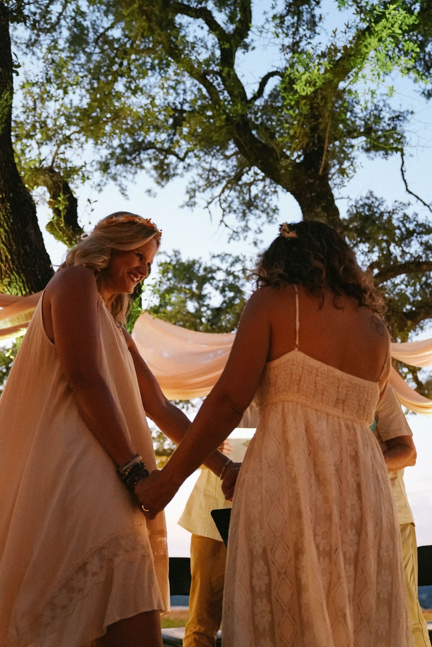 Two brides in white dresses hold hands during an outdoor wedding ceremony under a large oak tree.