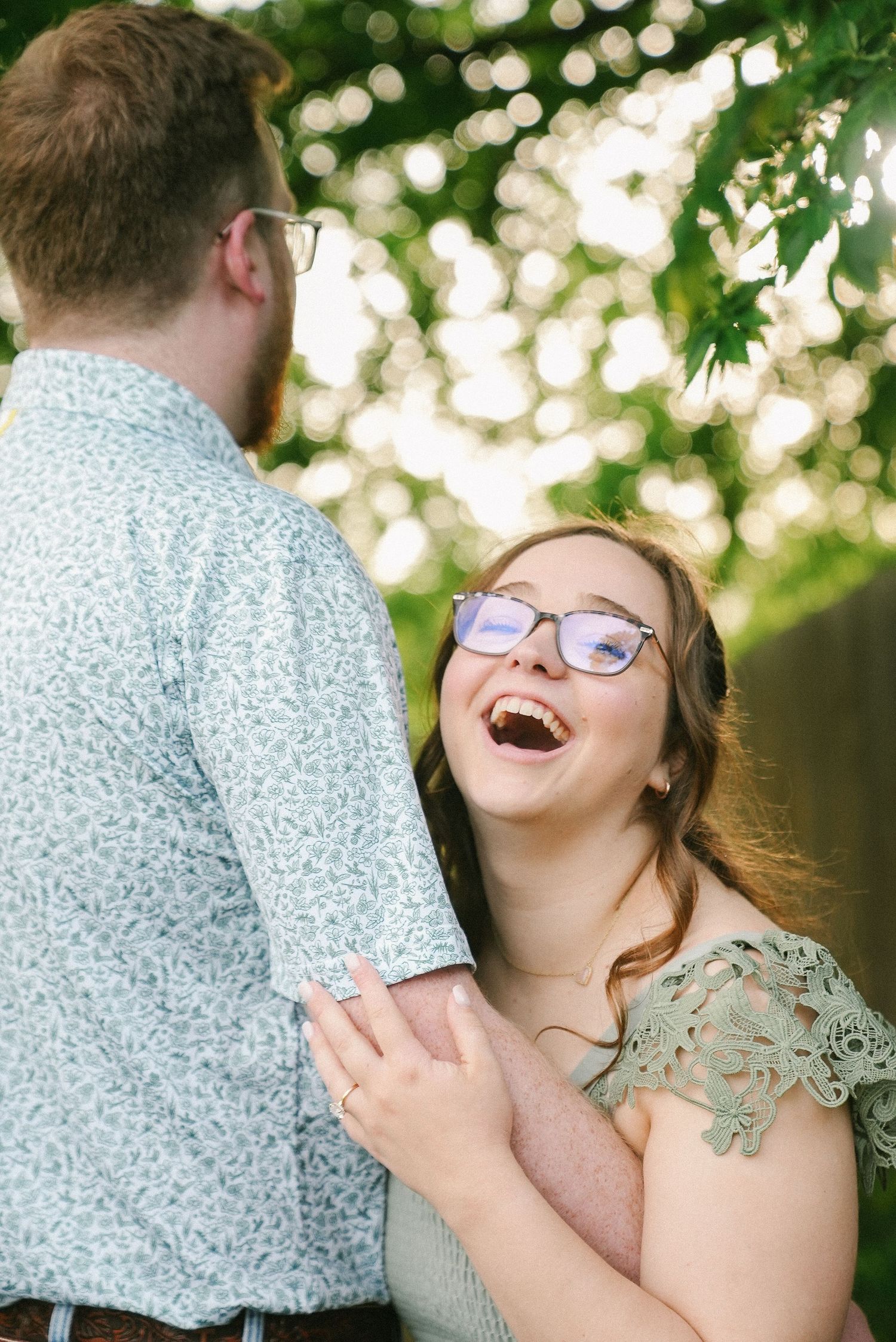Joyful woman with glasses laughing while holding hands with partner in sunlit garden setting.