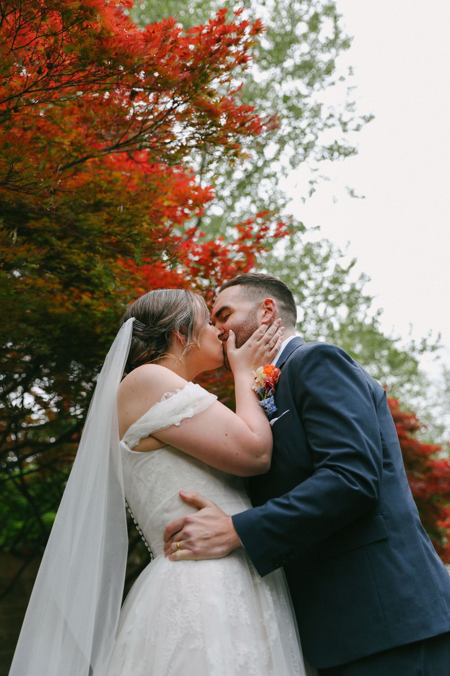 Bride and groom share romantic kiss beneath striking red maple trees in garden setting.