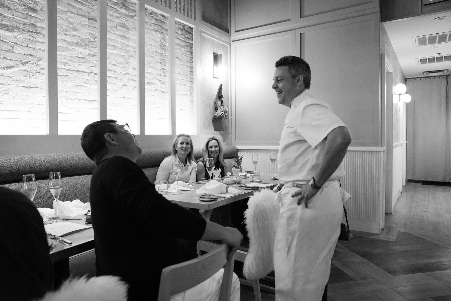 Black and white photo of a chef speaking with guests at a restaurant table.