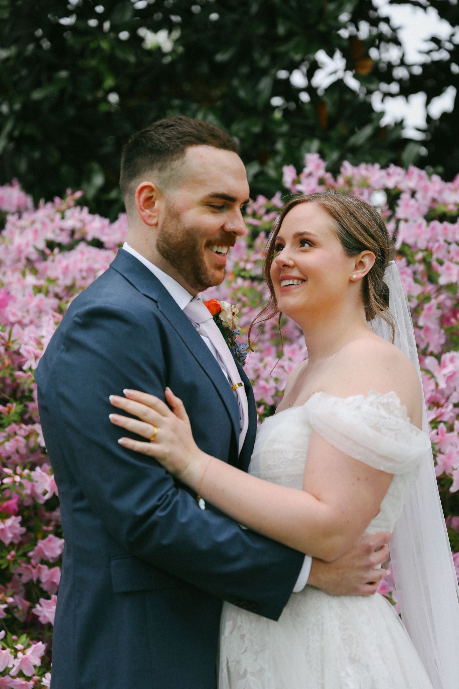 Smiling bride and groom embracing in front of vibrant pink flowering bushes outdoors.