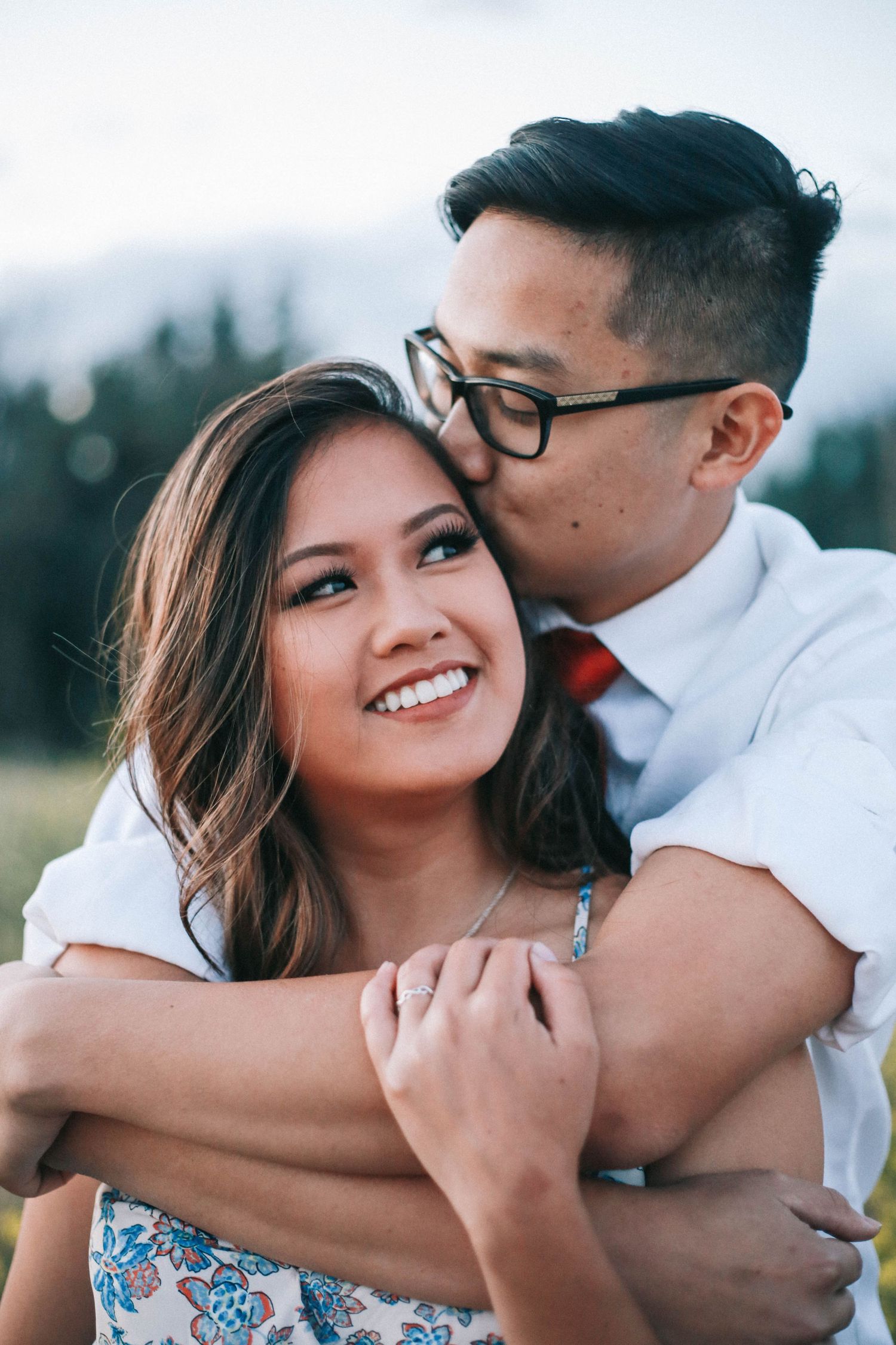 A man wearing glasses embraces and kisses a smiling woman on the cheek in an outdoor field setting.