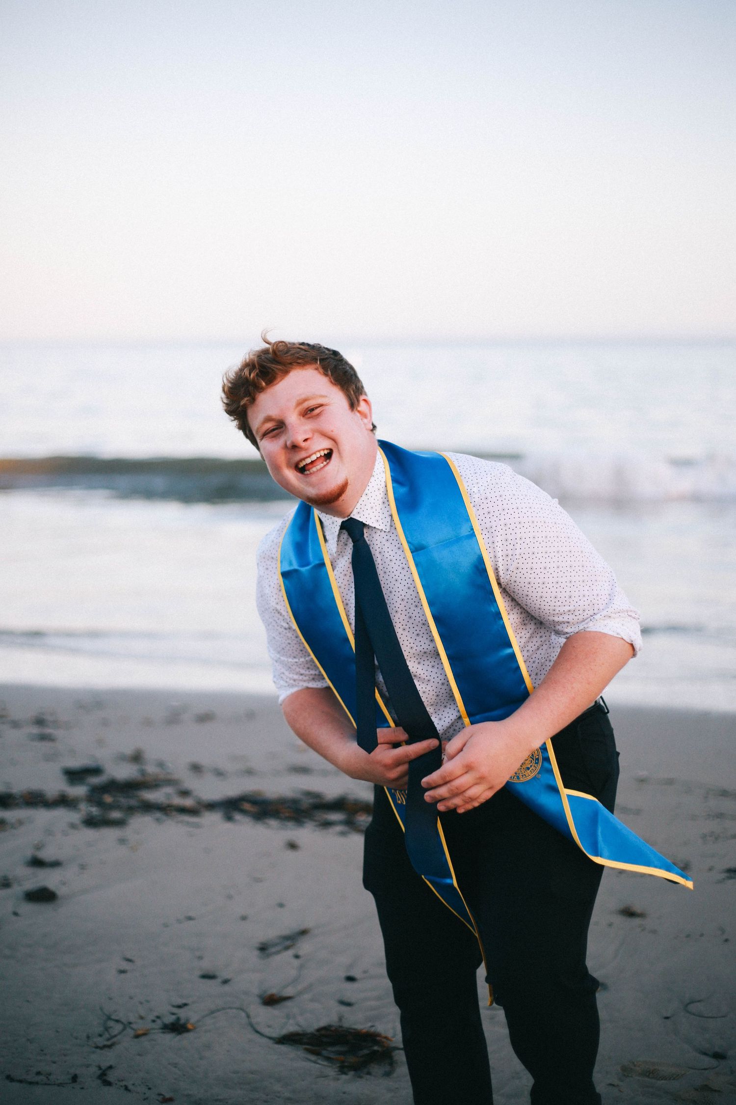 Laughing young man in white shirt with blue graduation stole stands on a sandy beach.