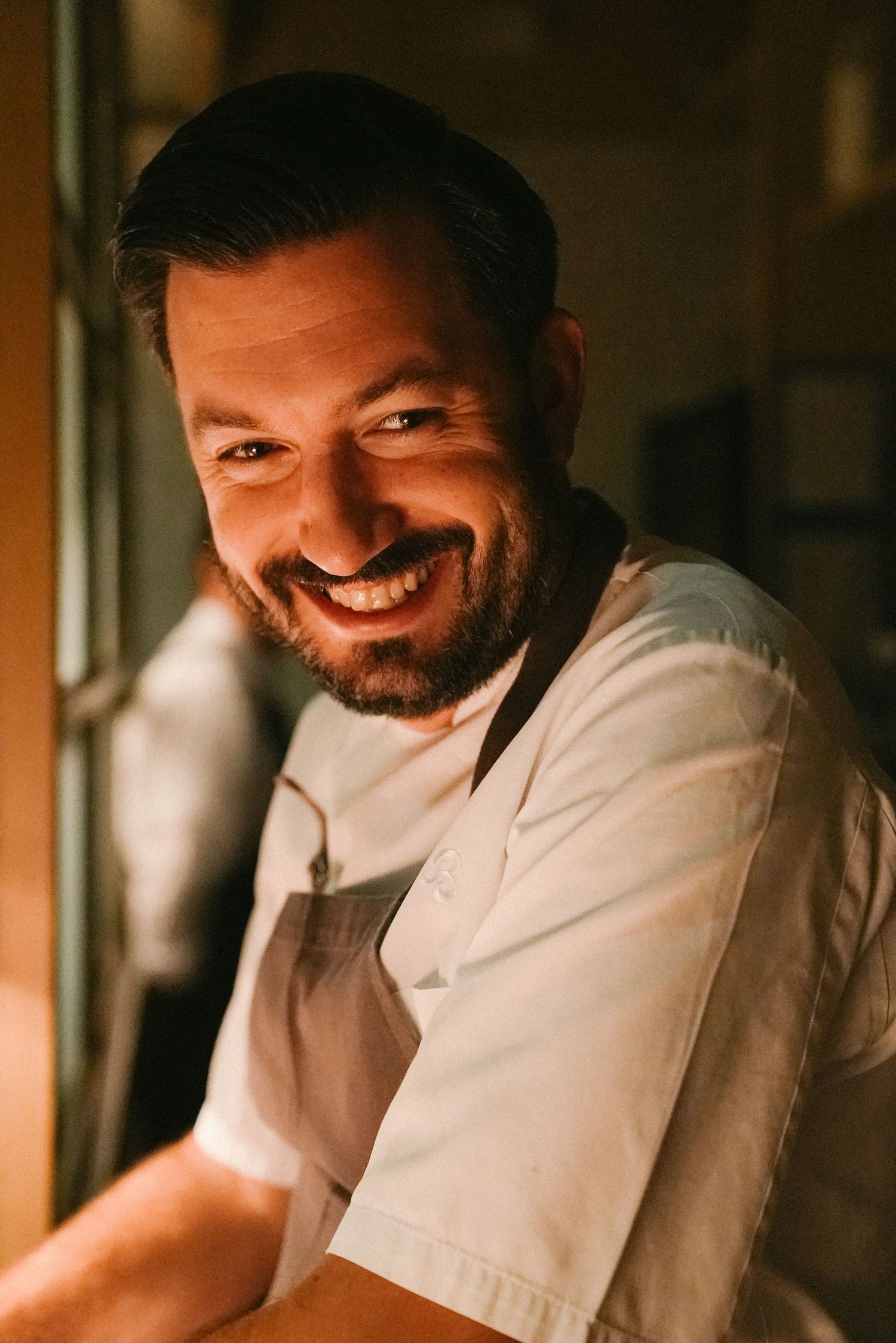 Smiling male chef in white apron and jacket poses warmly in a restaurant kitchen doorway.